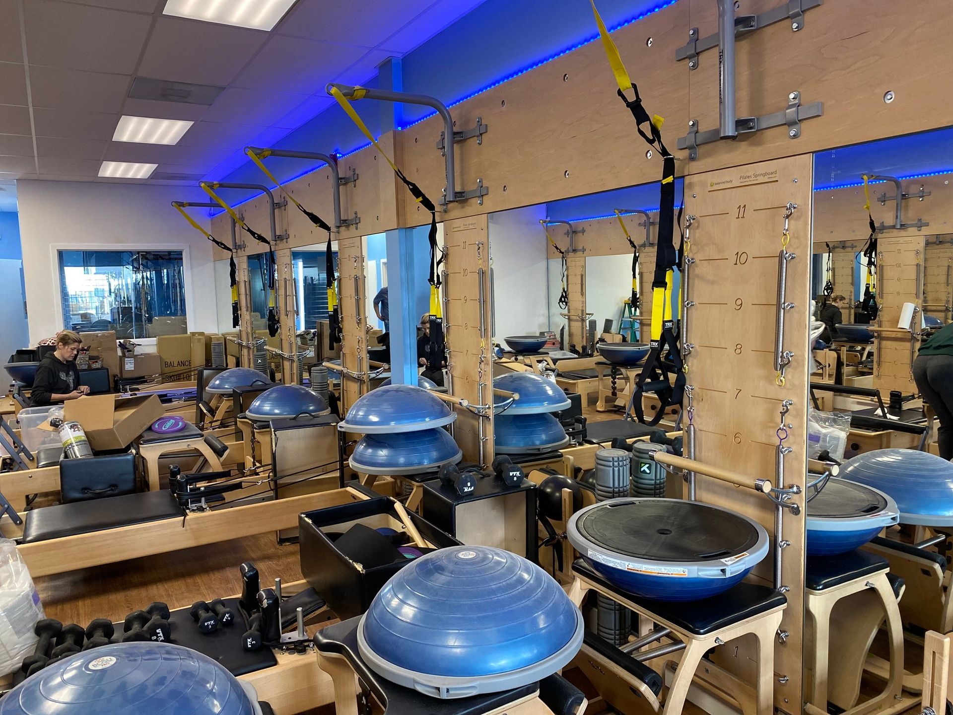 A gym interior filled with exercise equipment, including BOSU balls, pilates reformers, and TRX straps hanging on walls.