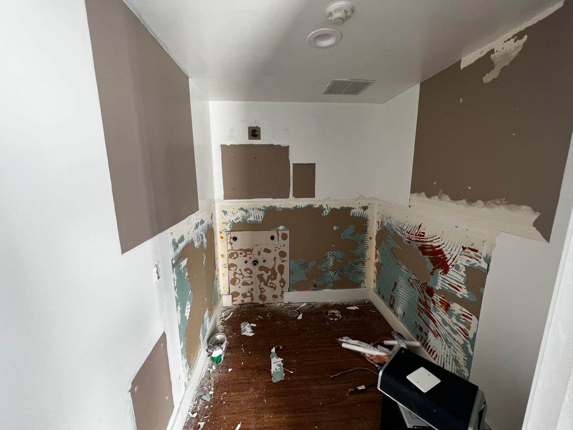 A small room undergoing renovations with exposed drywall, patched walls, and construction debris on the wooden floor.