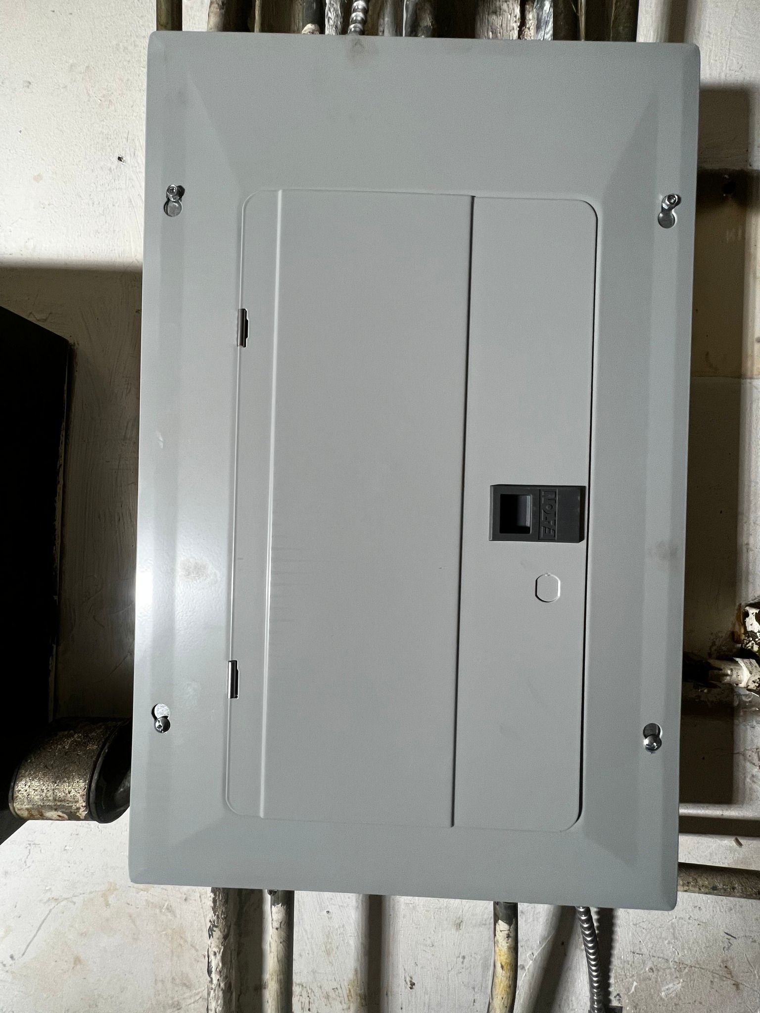 A gray electrical service panel box mounted on a wall with conduits visible at the top and bottom.