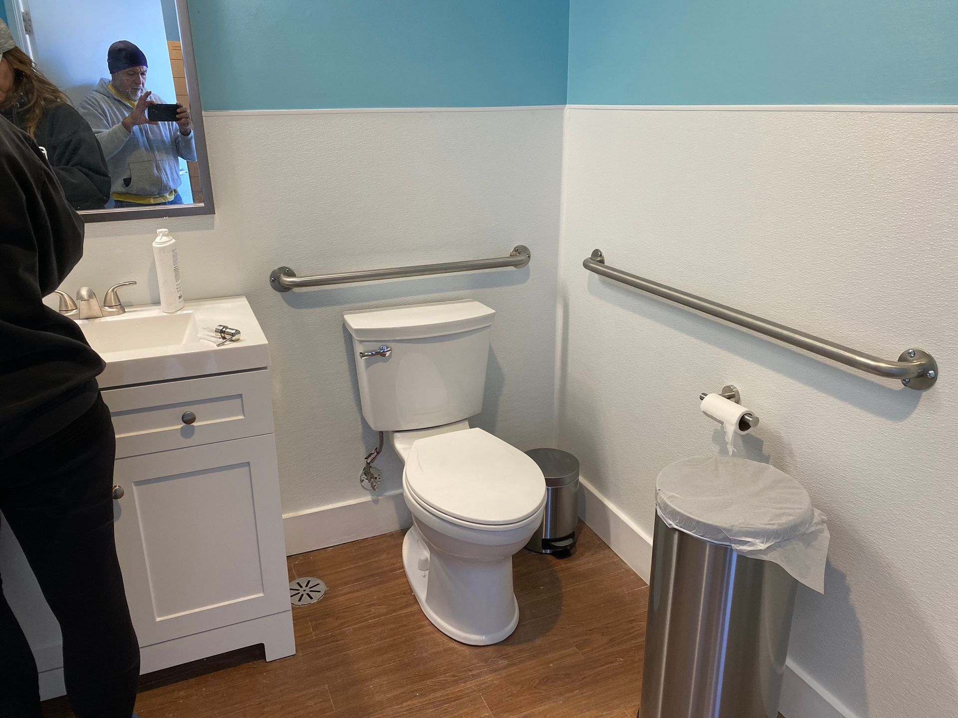 Accessible bathroom with a toilet, grab bars, sink, mirror, and trash bin against light blue and white painted walls.