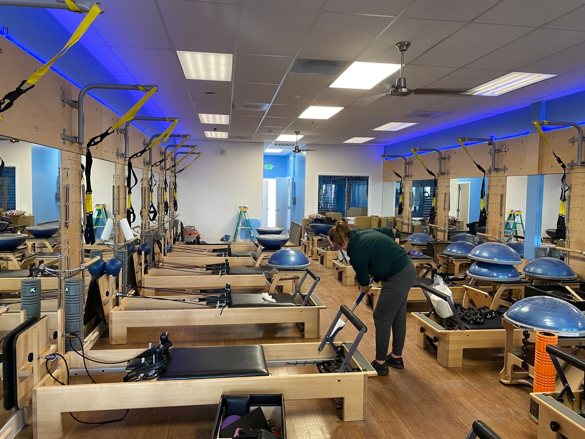A studio with rows of Pilates reformers and blue balance trainers as a person cleans the hardwood floor.