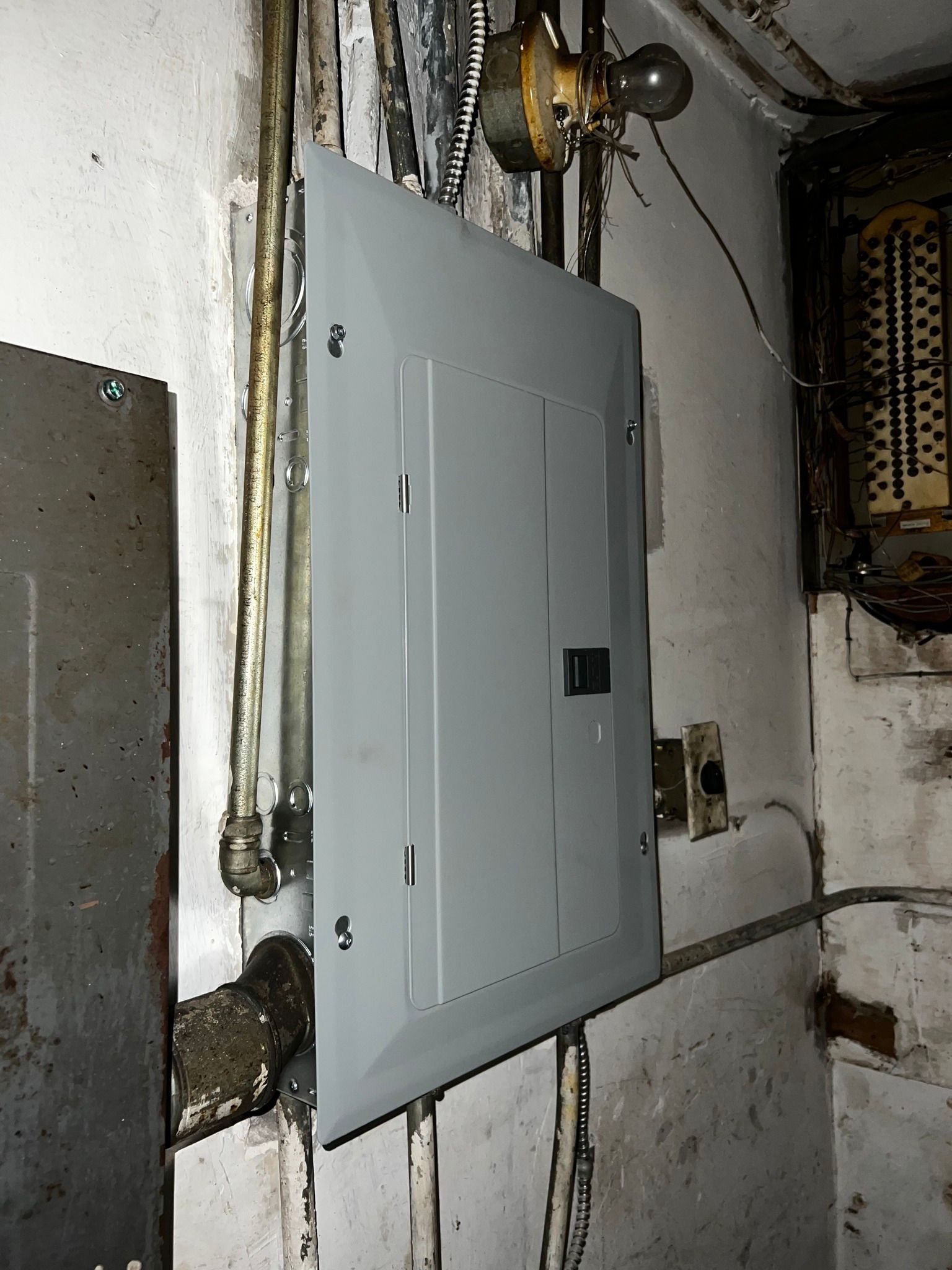 A gray electrical service panel mounted on a wall in a basement next to an exposed, older breaker box and a bare lightbulb.