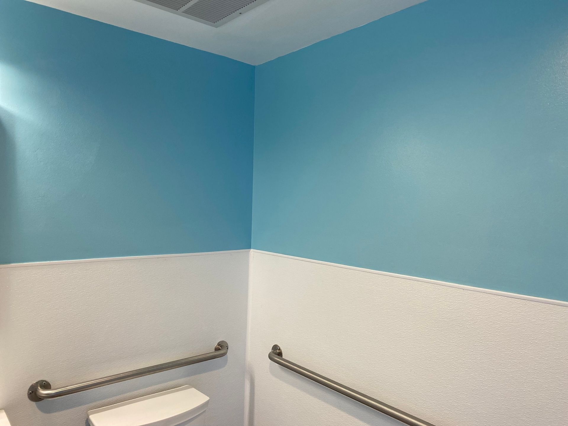 A bathroom corner with blue upper walls, white textured lower walls, and silver horizontal grab bars.