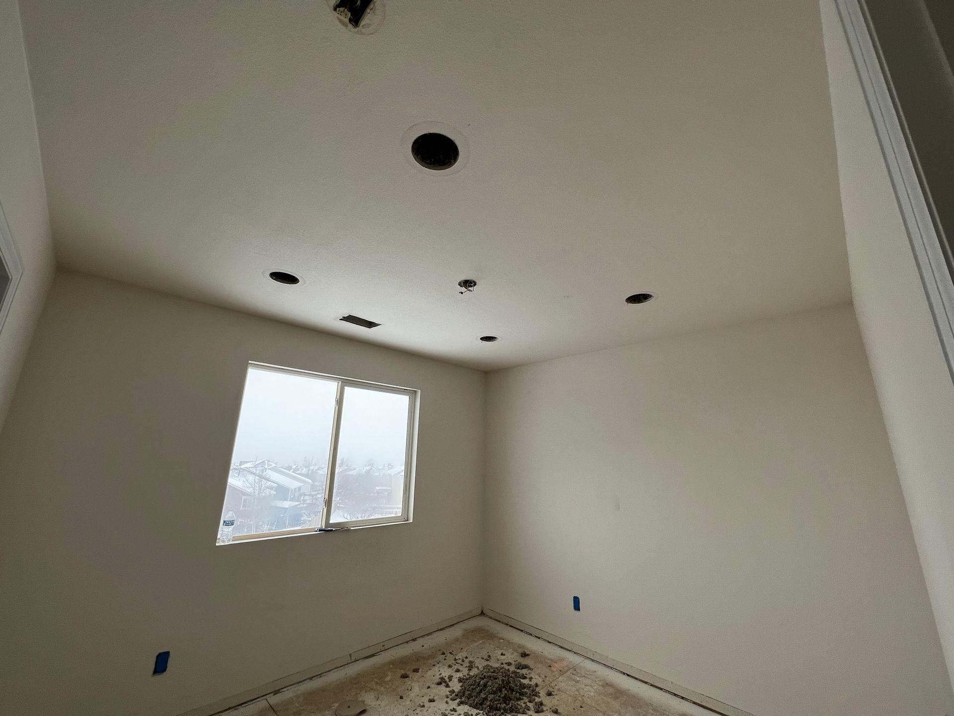An unfinished room with light-colored walls, a window, and multiple holes cut into the ceiling for lighting fixtures.