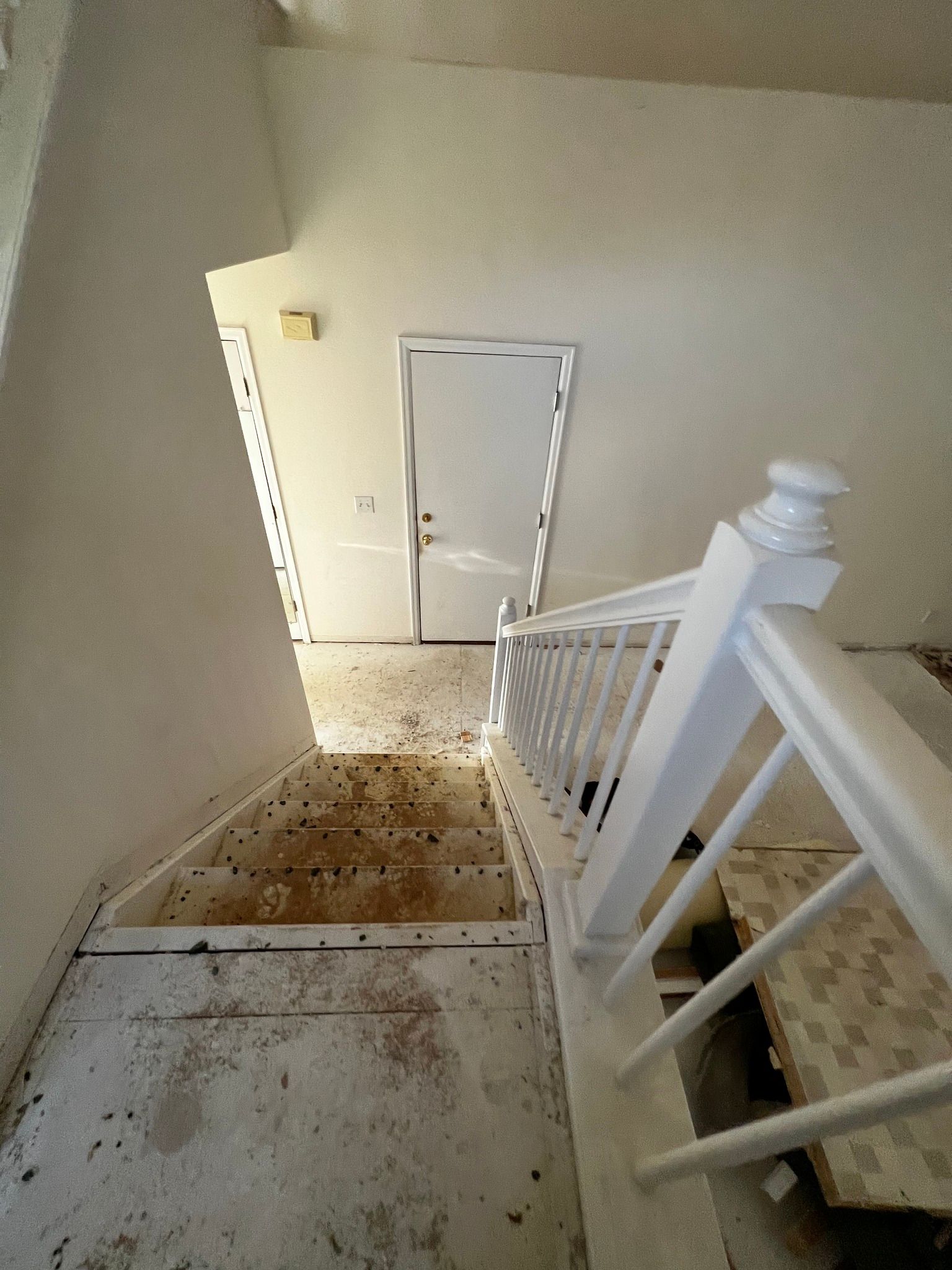 Top-down view of unfinished wooden stairs with a white railing leading to a white door in a sparse, beige-walled hallway.
