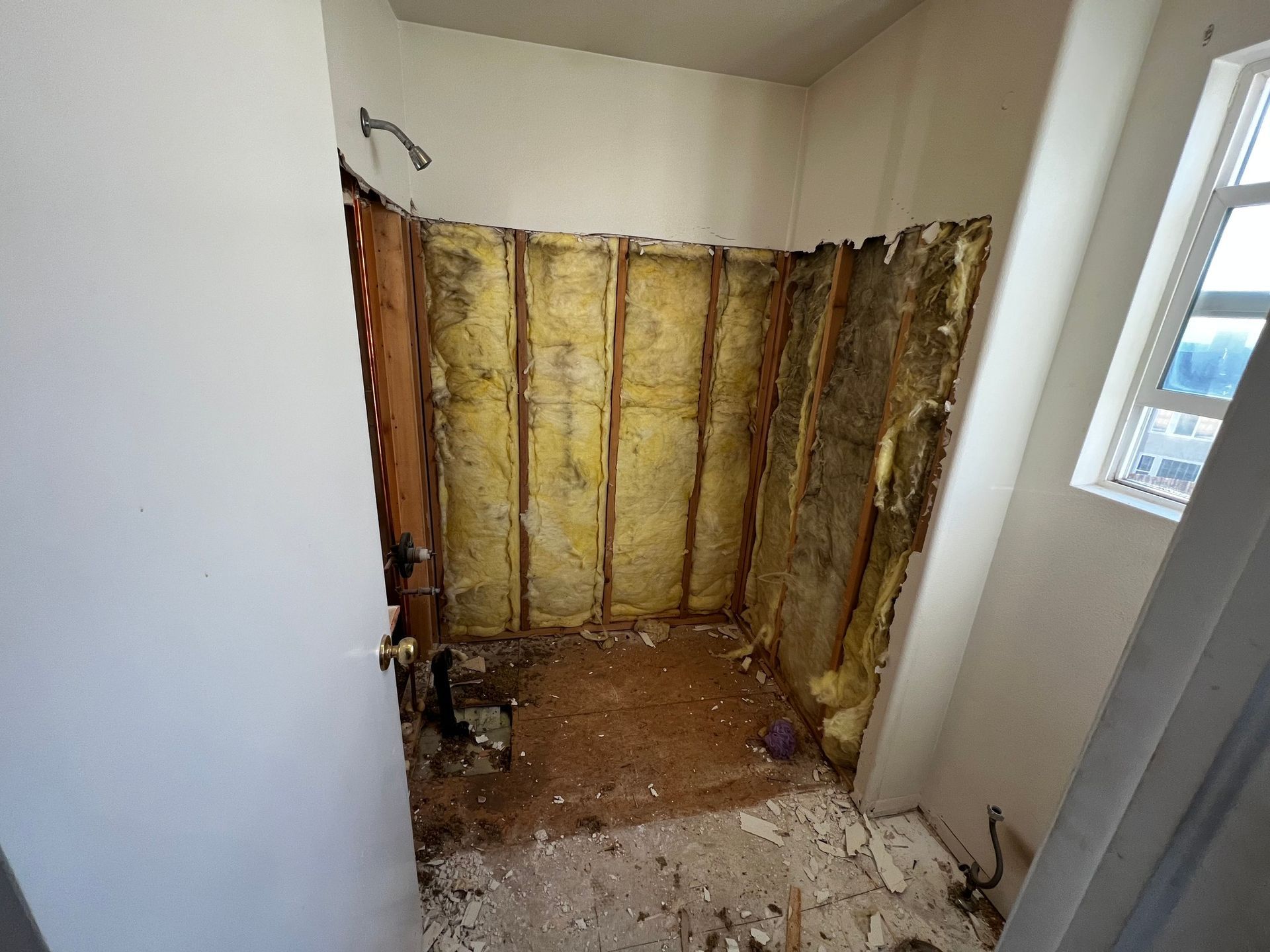A bathroom under renovation with exposed wood wall studs, yellow insulation, and debris on the floor.