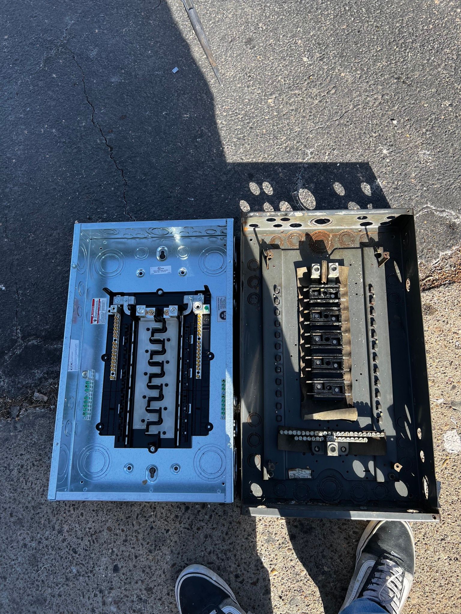 Two electrical panel interiors, one new and silver and one used and black, resting on asphalt pavement.