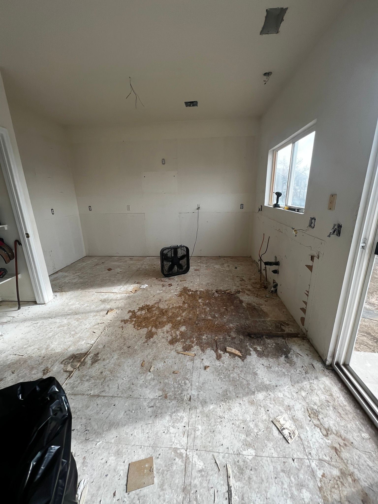 A room under renovation with bare drywall, unfinished concrete flooring, a floor fan in the center, and a large window.