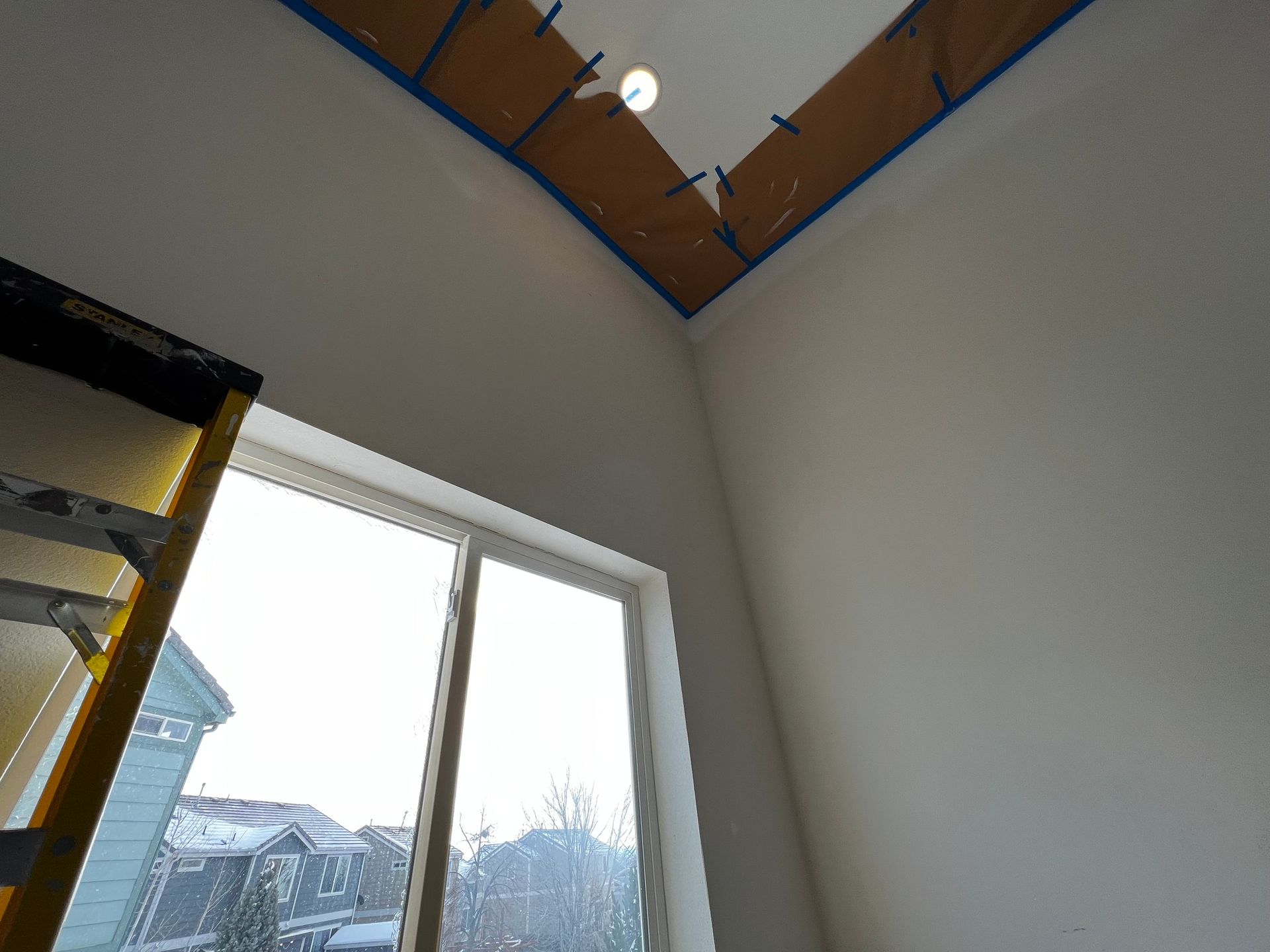 A partially painted high ceiling in a room with a large window and a ladder in the foreground.