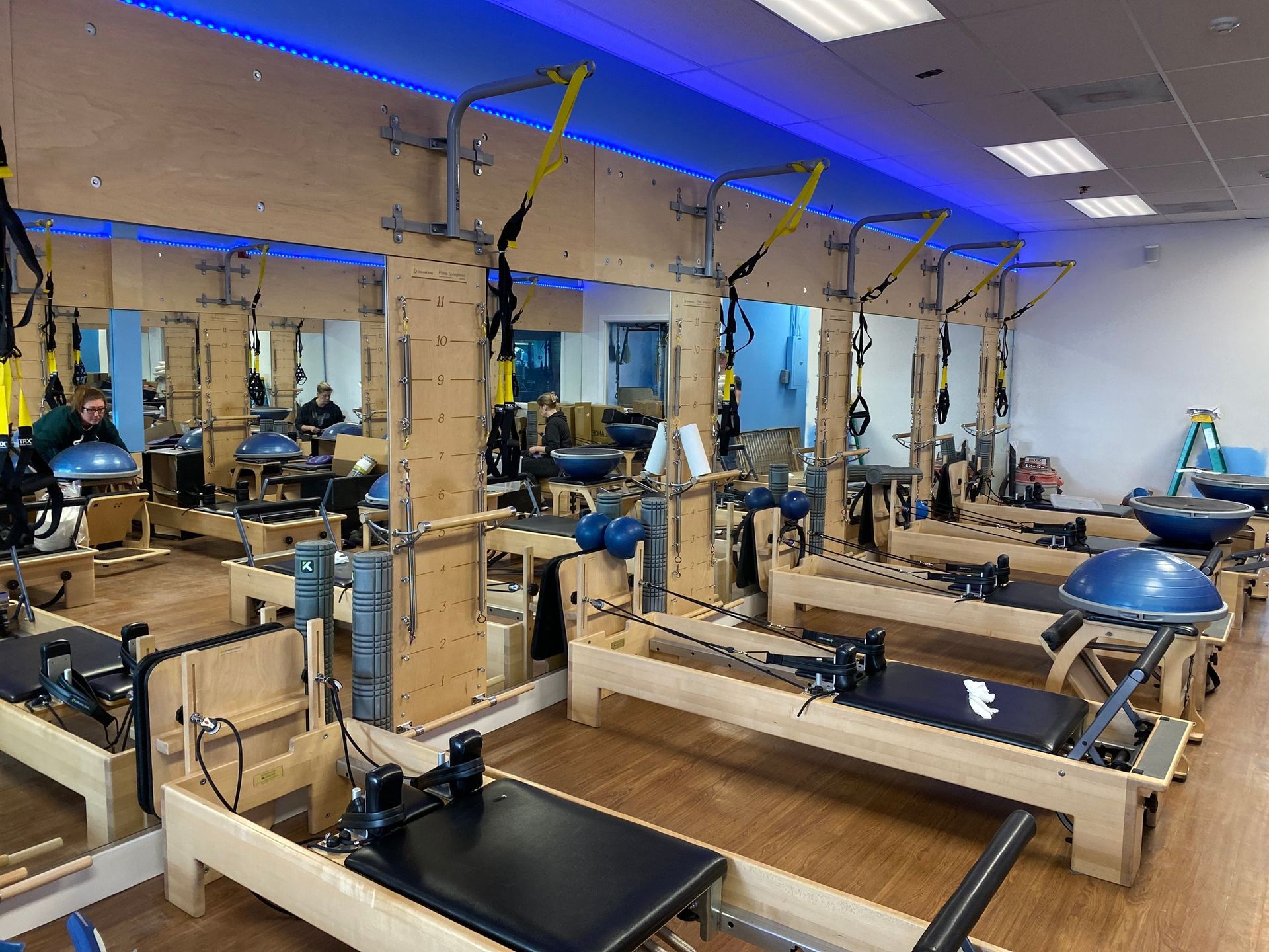 A fitness studio lined with Pilates reformers and TRX suspension trainers, featuring mirrors and blue accent lighting.