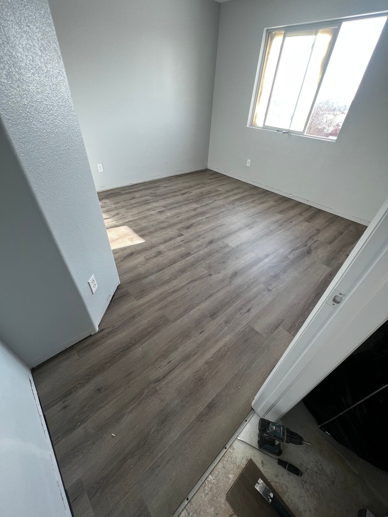 An empty room with gray walls and wood-look laminate flooring, seen from a doorway with construction tools nearby.