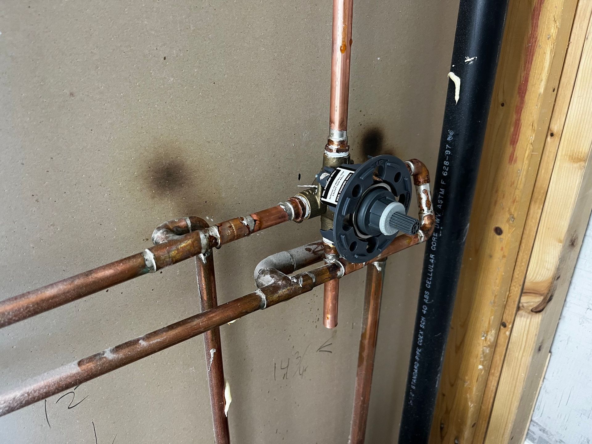A plumbing shower mixing valve installed on copper pipes against a wall with a black pipe to the side.