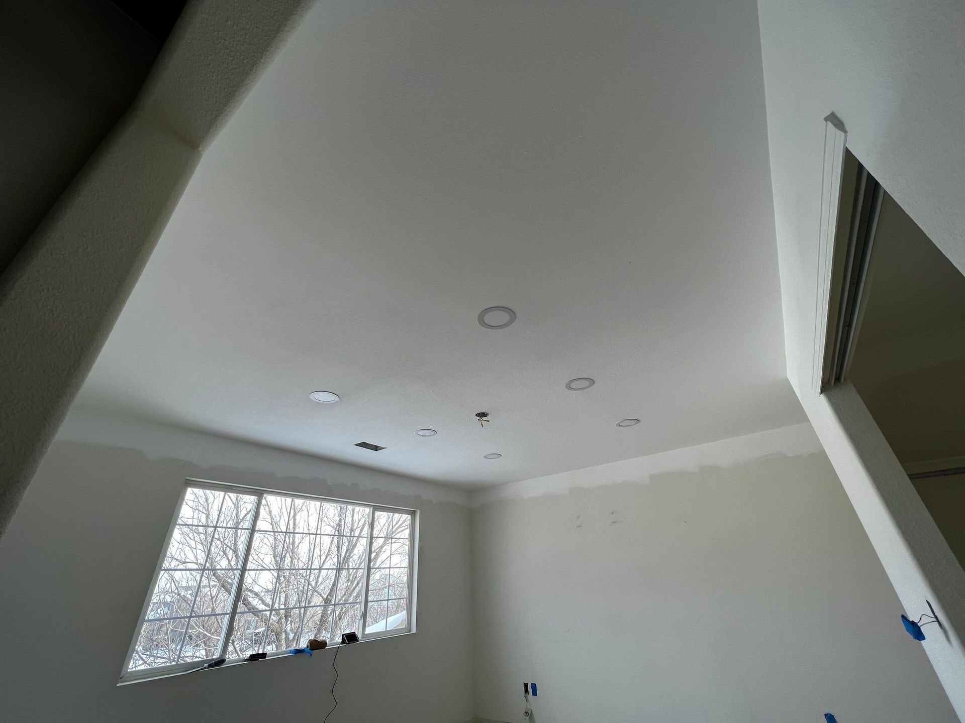 An unfinished room with white drywall on walls and ceiling, a window view of trees, and recessed lighting fixtures.