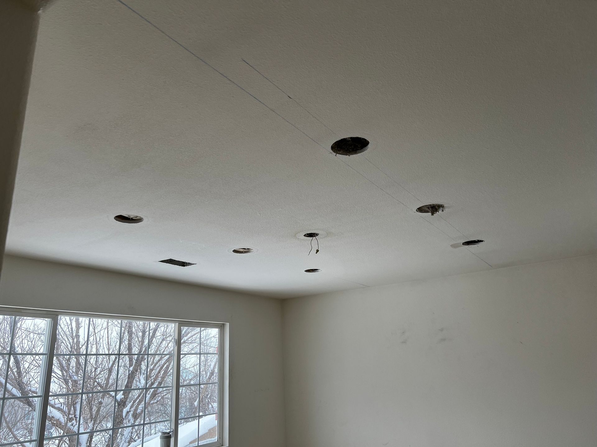 A room ceiling with a textured finish, featuring several circular holes for recessed lighting and wires exposed.