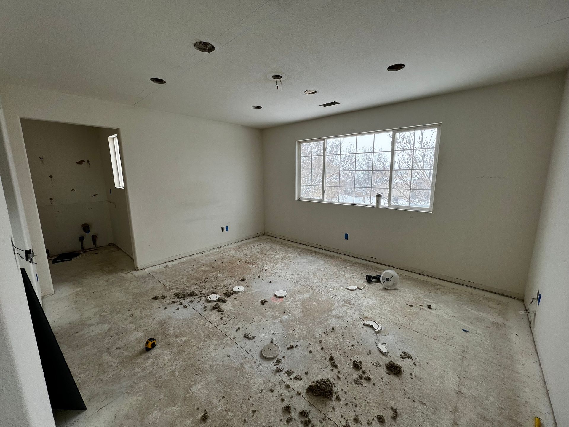 Empty, cream-colored room under renovation with a large window, recessed lighting, and debris scattered on the floor.