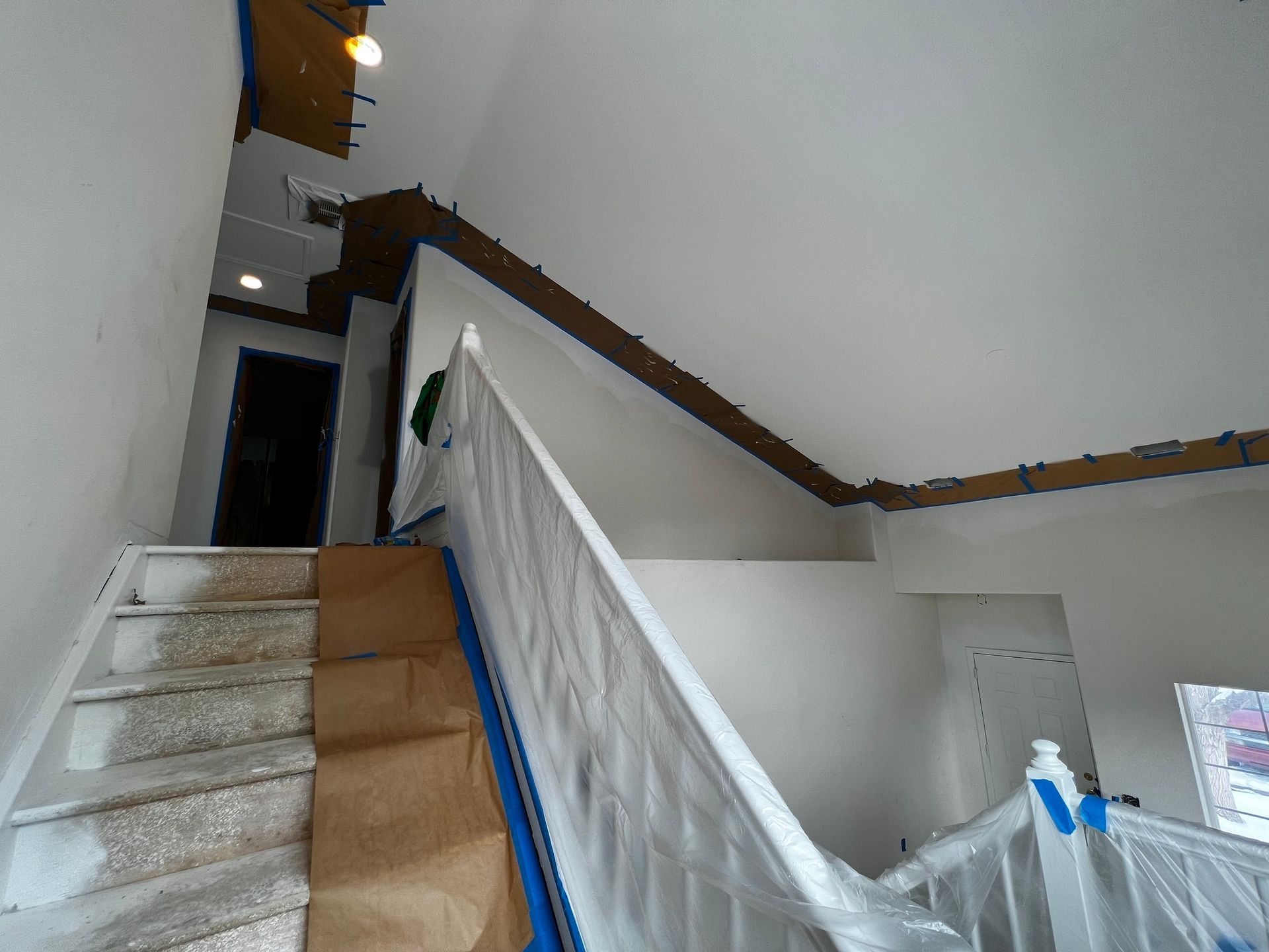 An interior view of a staircase undergoing painting, with blue tape, protective paper, and plastic sheets covering surfaces.