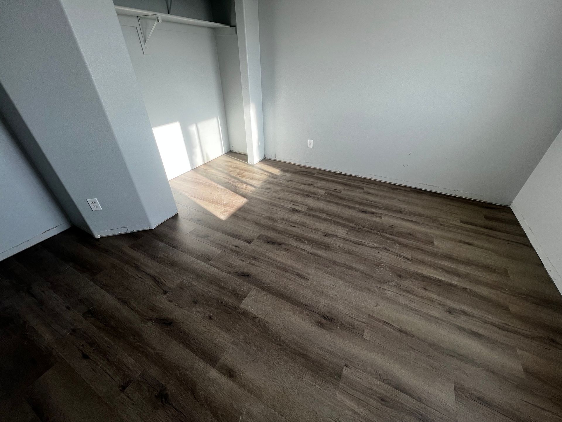 An empty, modern room with grey walls and dark wood-look laminate flooring, featuring a small alcove with a closet rail.