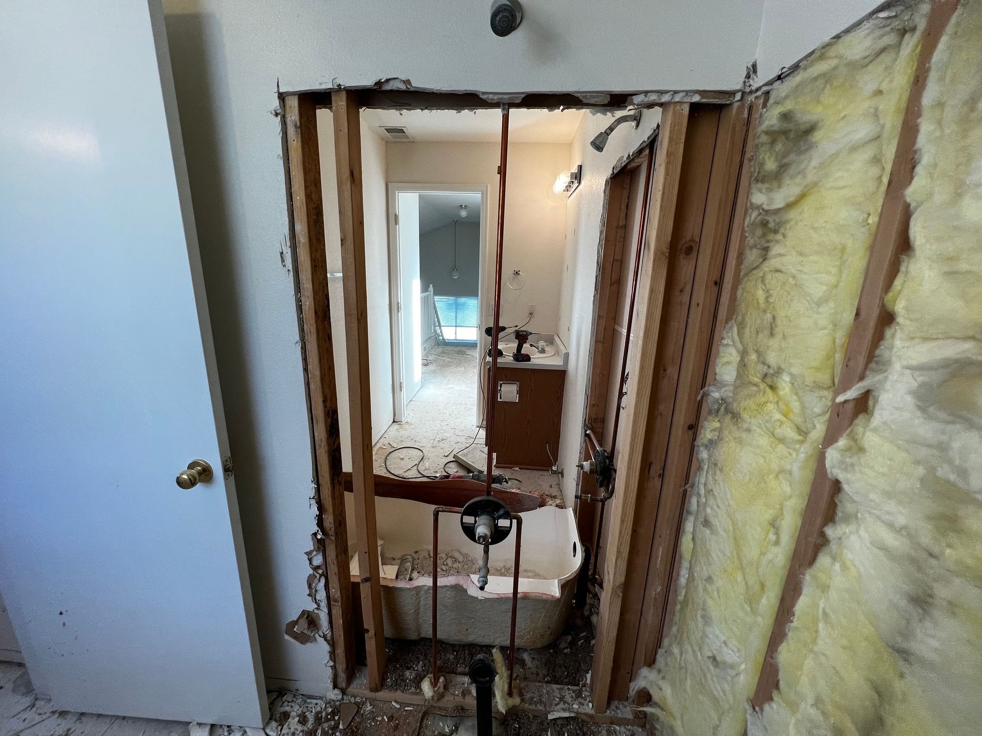 A bathroom undergoing renovation, showing exposed wall studs, yellow insulation, and plumbing fixtures in a framed doorway.