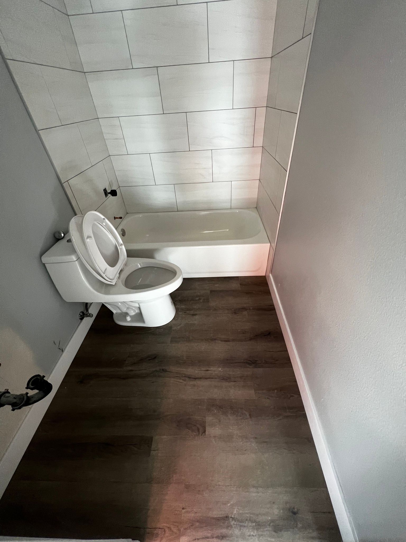 A small bathroom featuring dark wood flooring, light grey walls, a white toilet, and a white bathtub with tile surround.