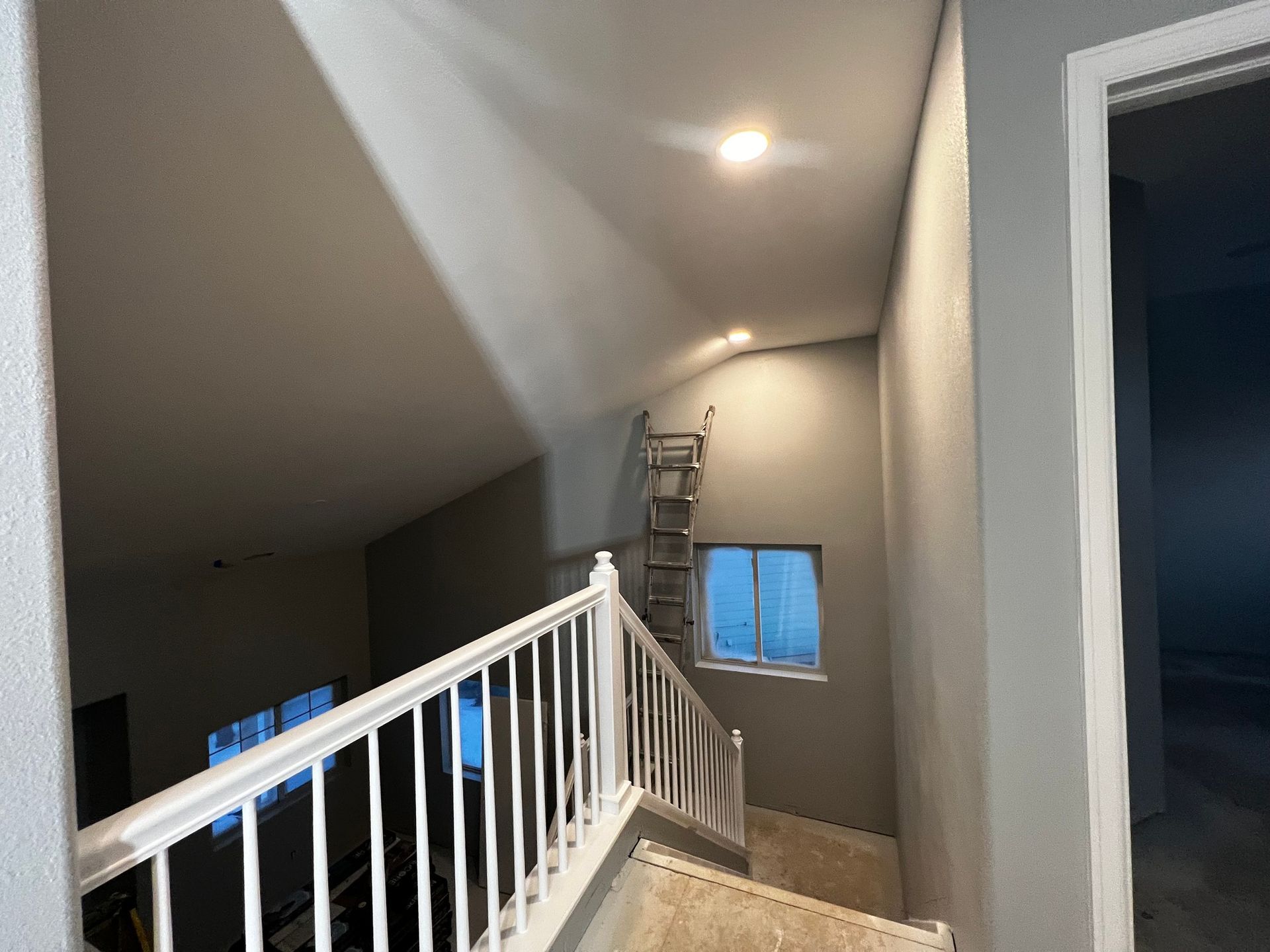 A staircase with a white railing leads up to a landing with a window, a metal ladder, and gray walls.