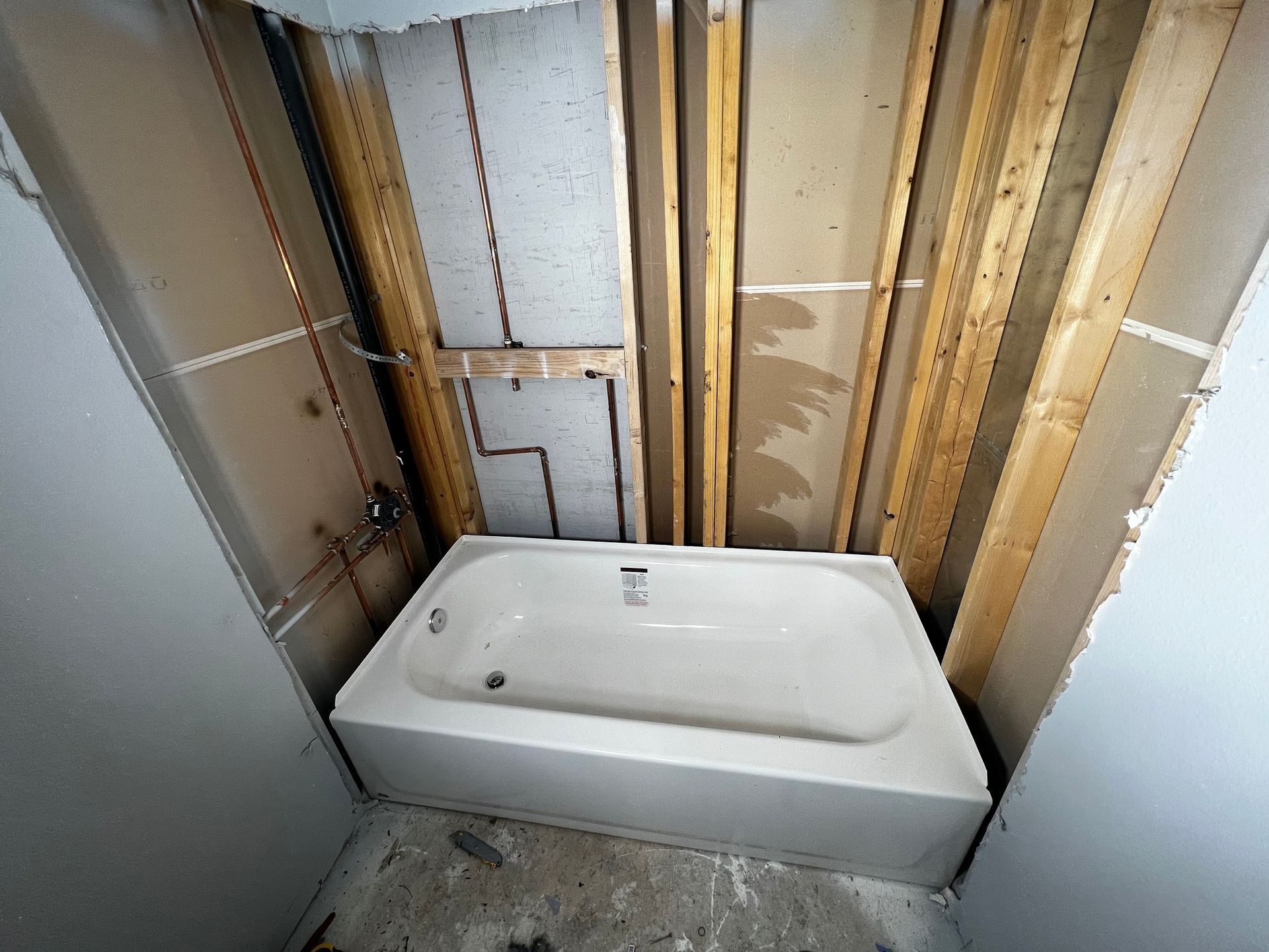 A white bathtub sits in an unfinished bathroom alcove with exposed wooden wall studs and copper plumbing pipes.
