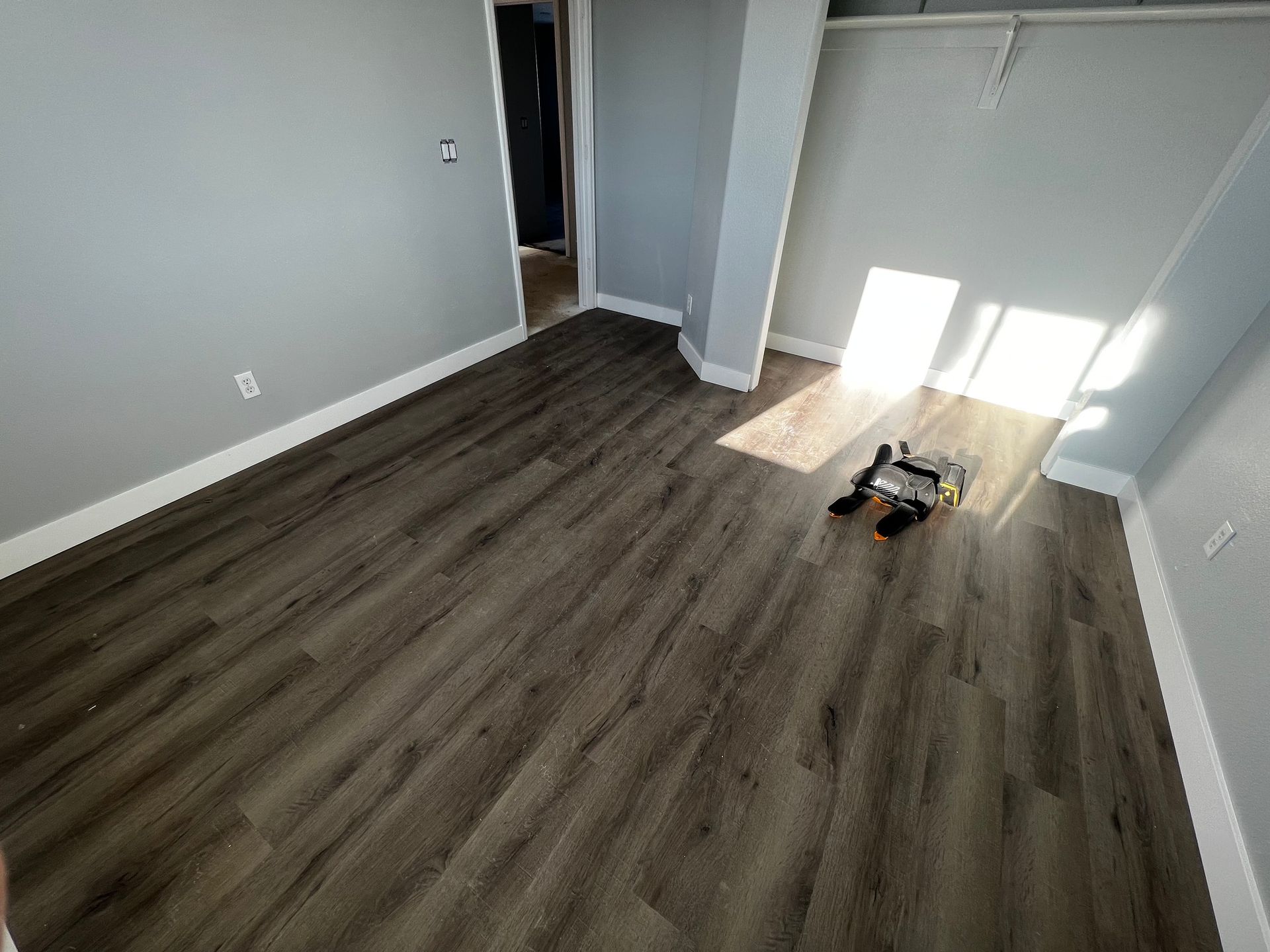 Empty room with grey walls, white trim, and dark wood-look flooring, featuring a small robotic vacuum on the floor.