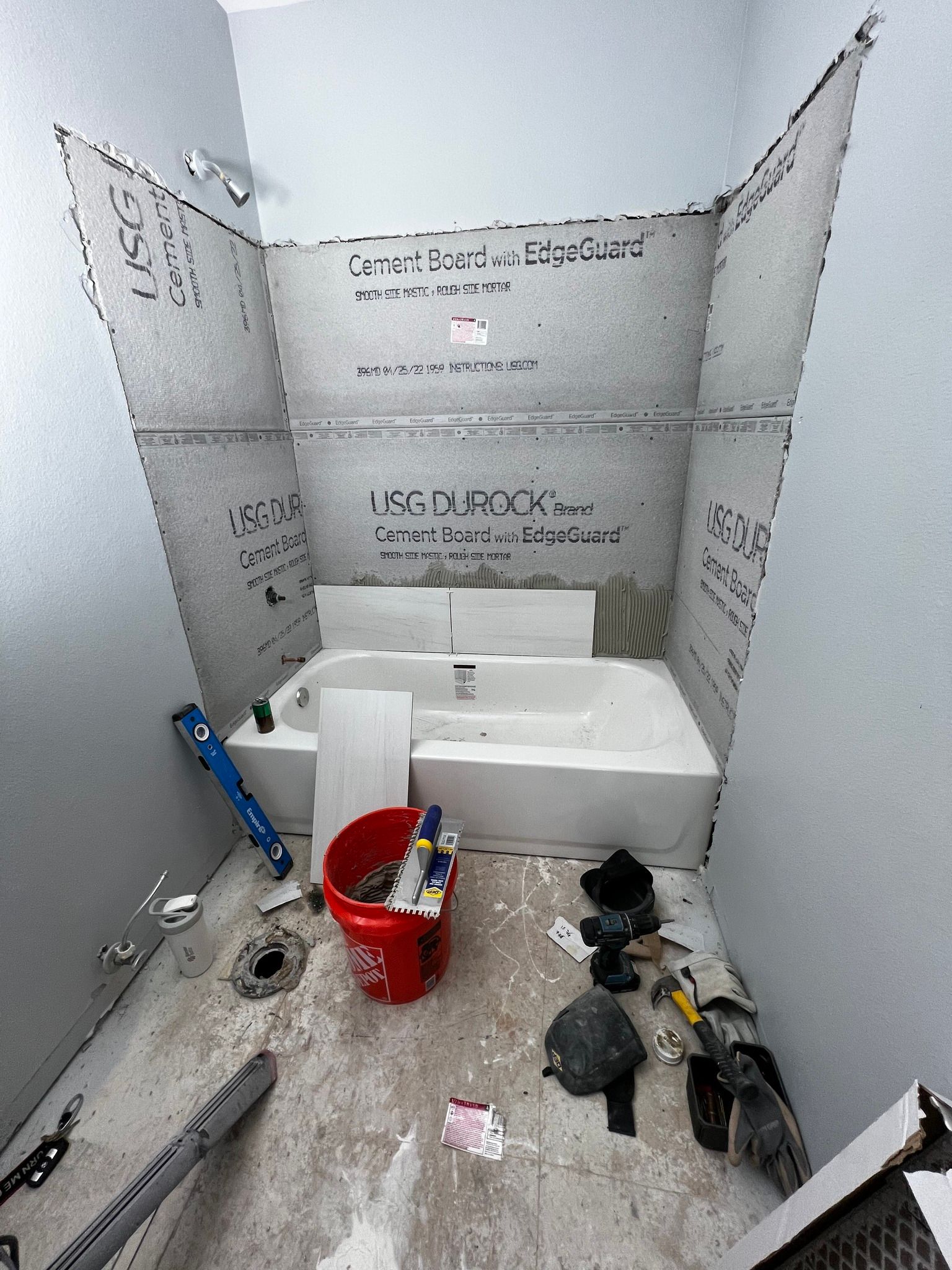 A partially installed shower surround with gray cement backer board, a white bathtub, and construction tools on the floor.