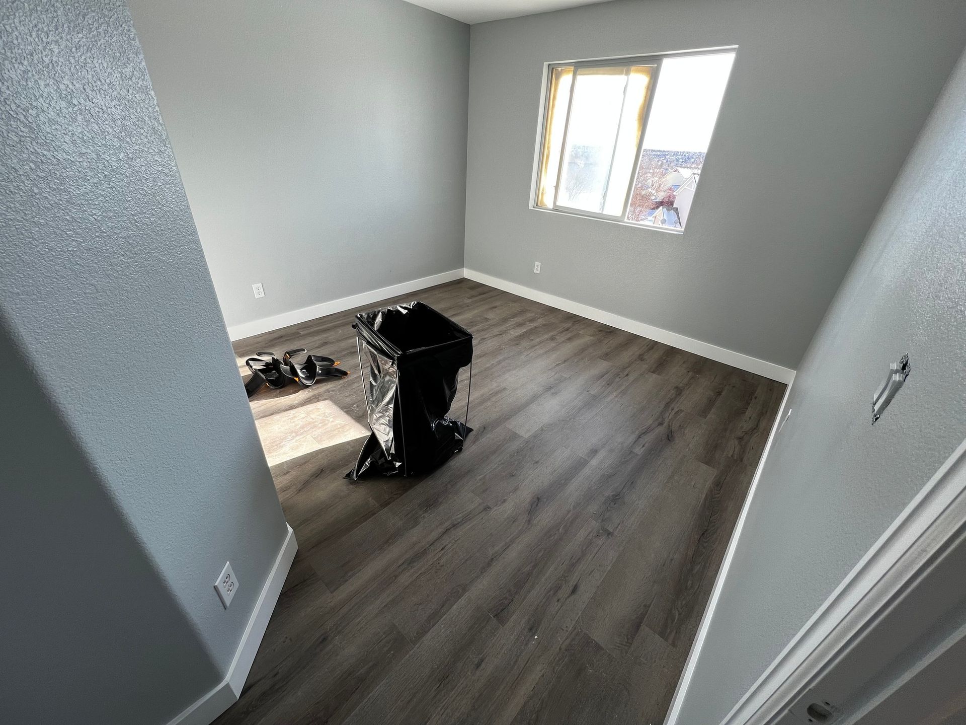 An empty room with gray walls, wood-look flooring, a window, and a black bassinet in the center.