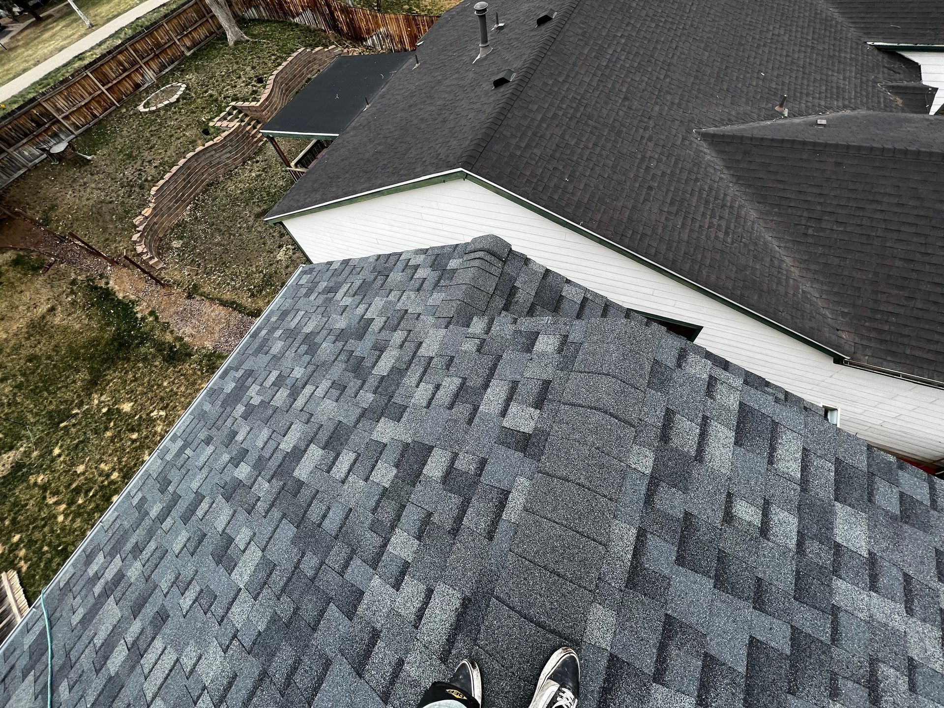 High-angle view of a grey shingled roof with a person's feet visible at the bottom edge, overlooking a backyard.