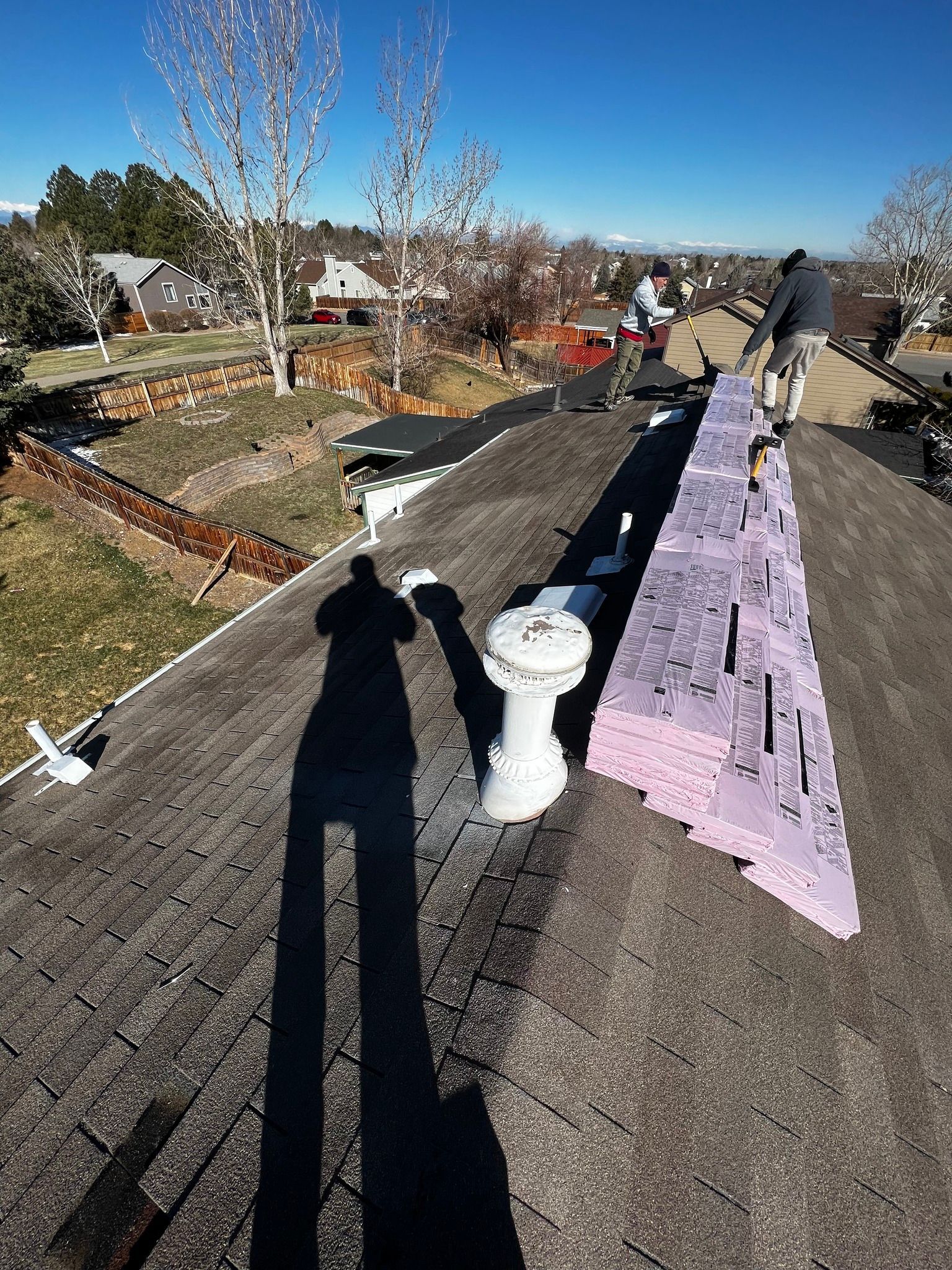 Two workers on a rooftop install pink foam insulation along the roof ridge under a clear blue sky.