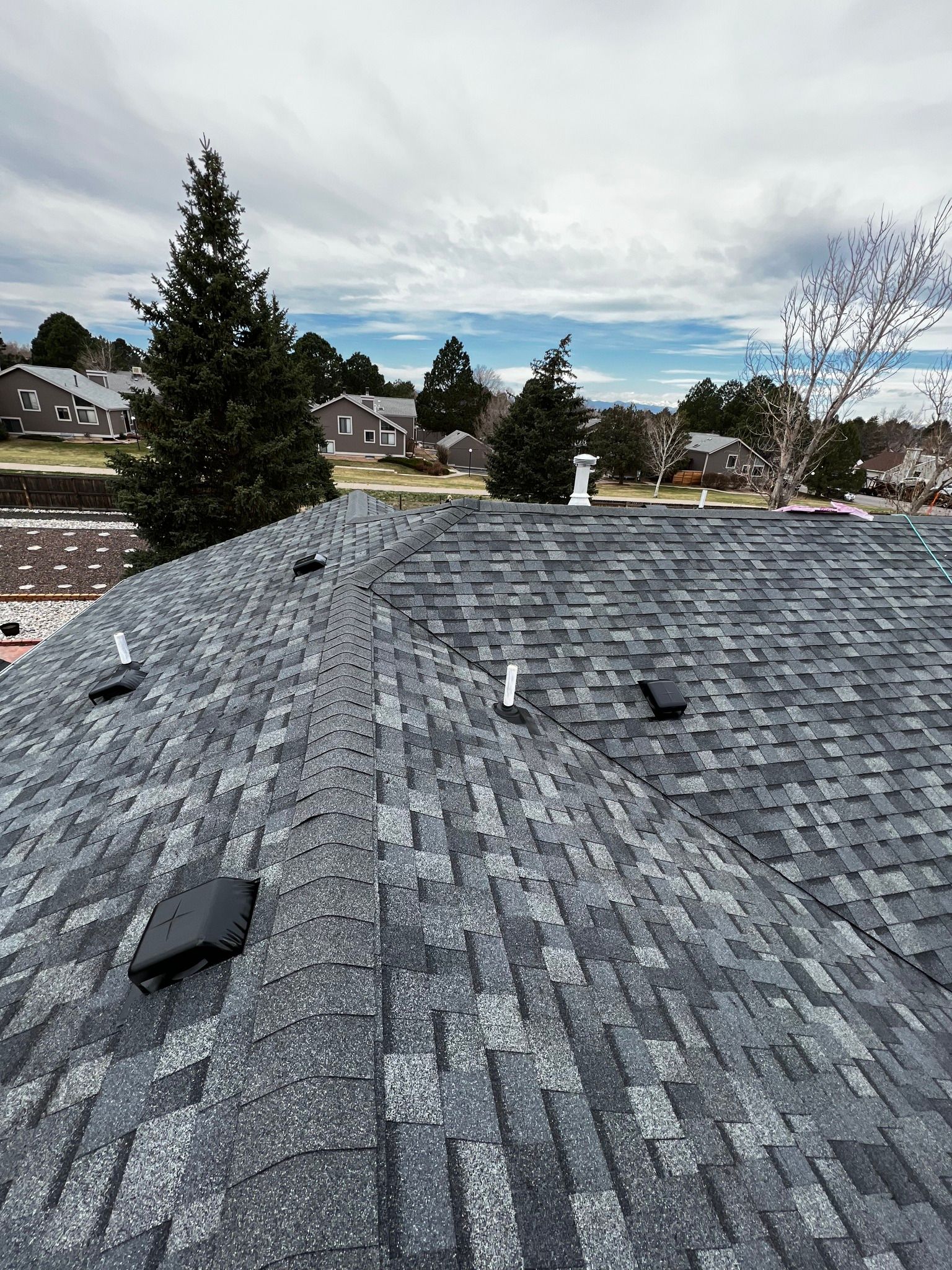An elevated view of a gray asphalt shingle roof featuring several vent pipes and square vents against a cloudy sky.