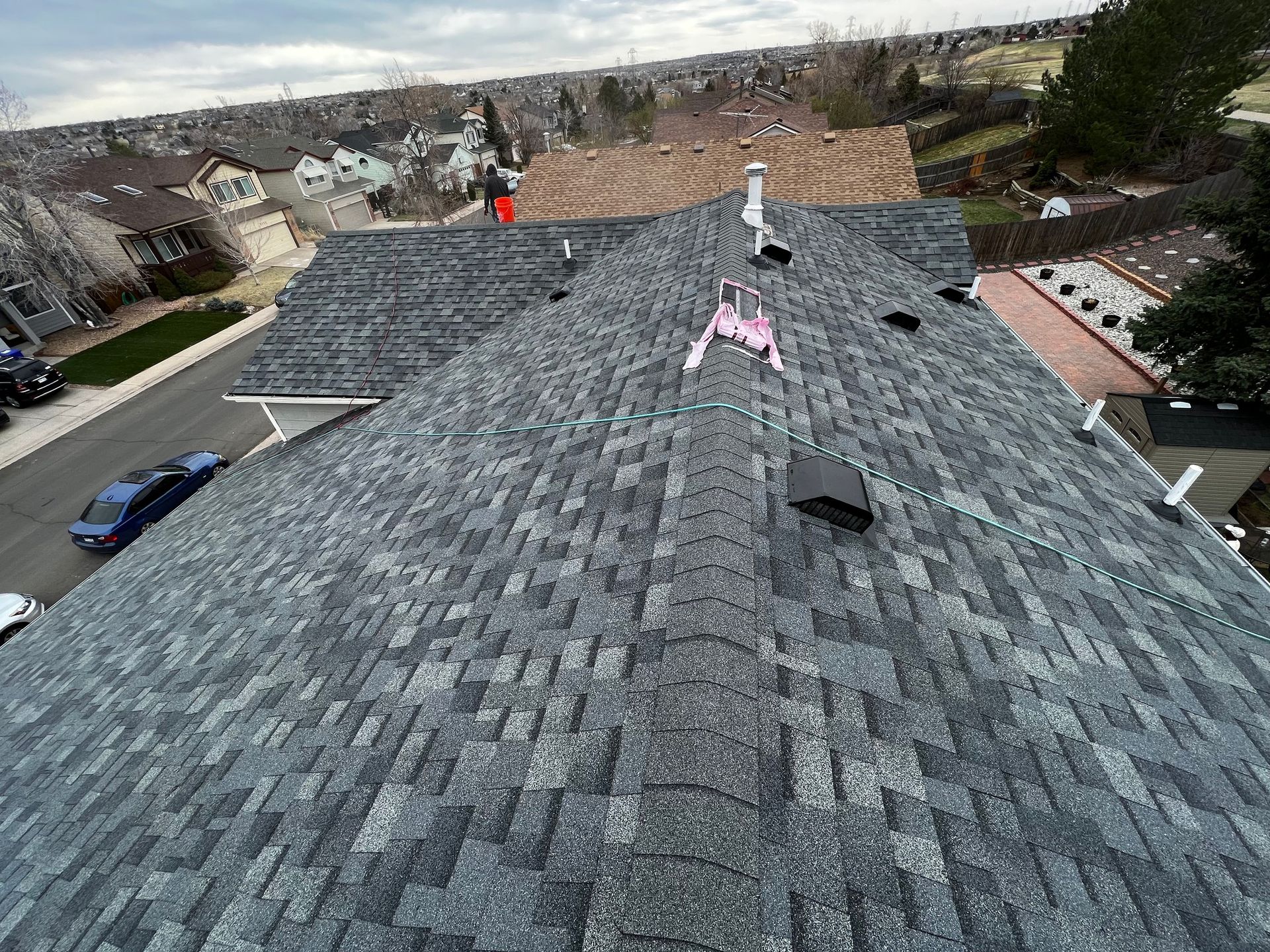 High-angle view of a grey shingled residential roof with several vents and a chimney against a suburban backdrop.