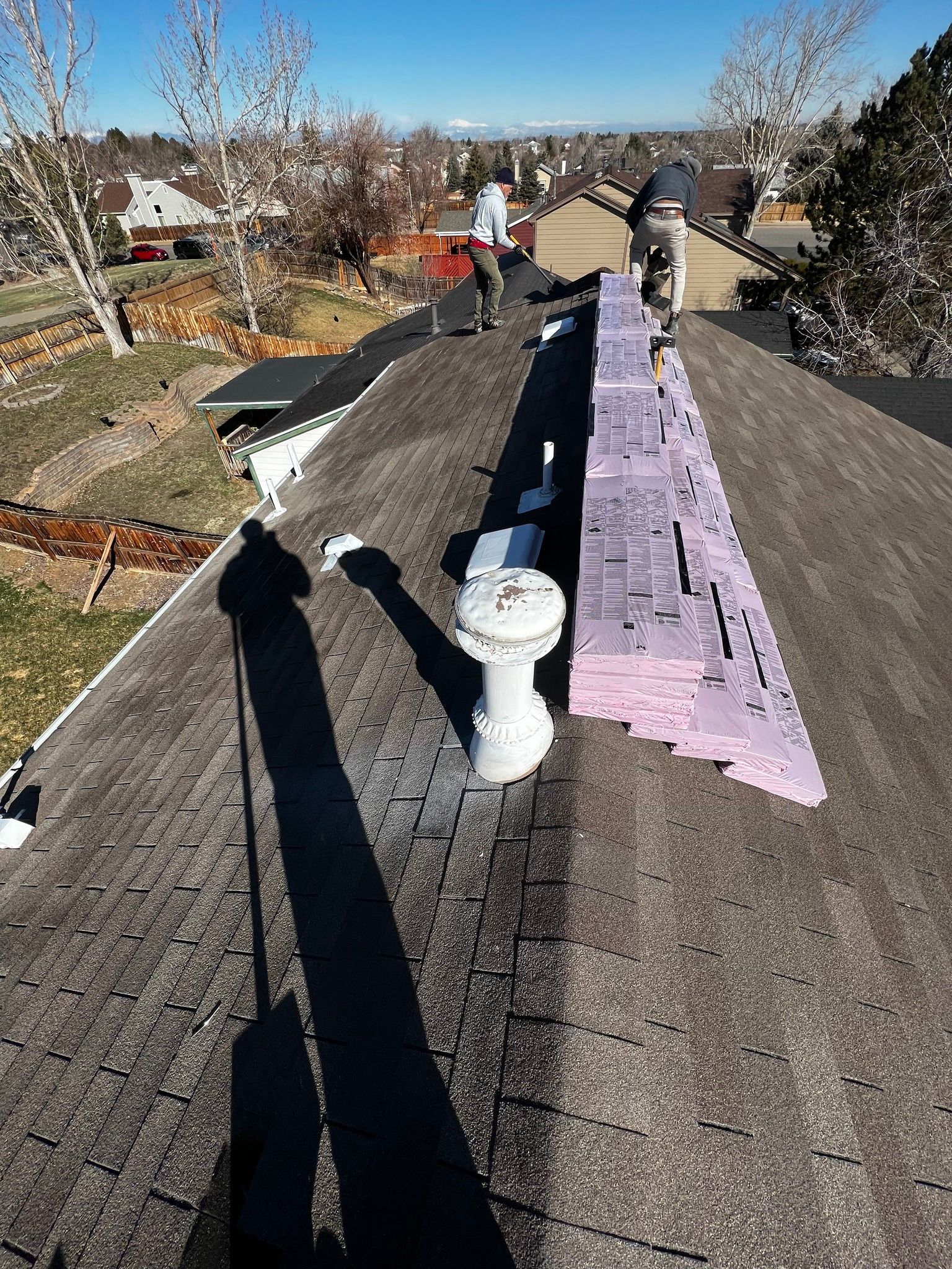 Two workers install purple foam insulation panels along the ridge of a residential roof on a sunny day.