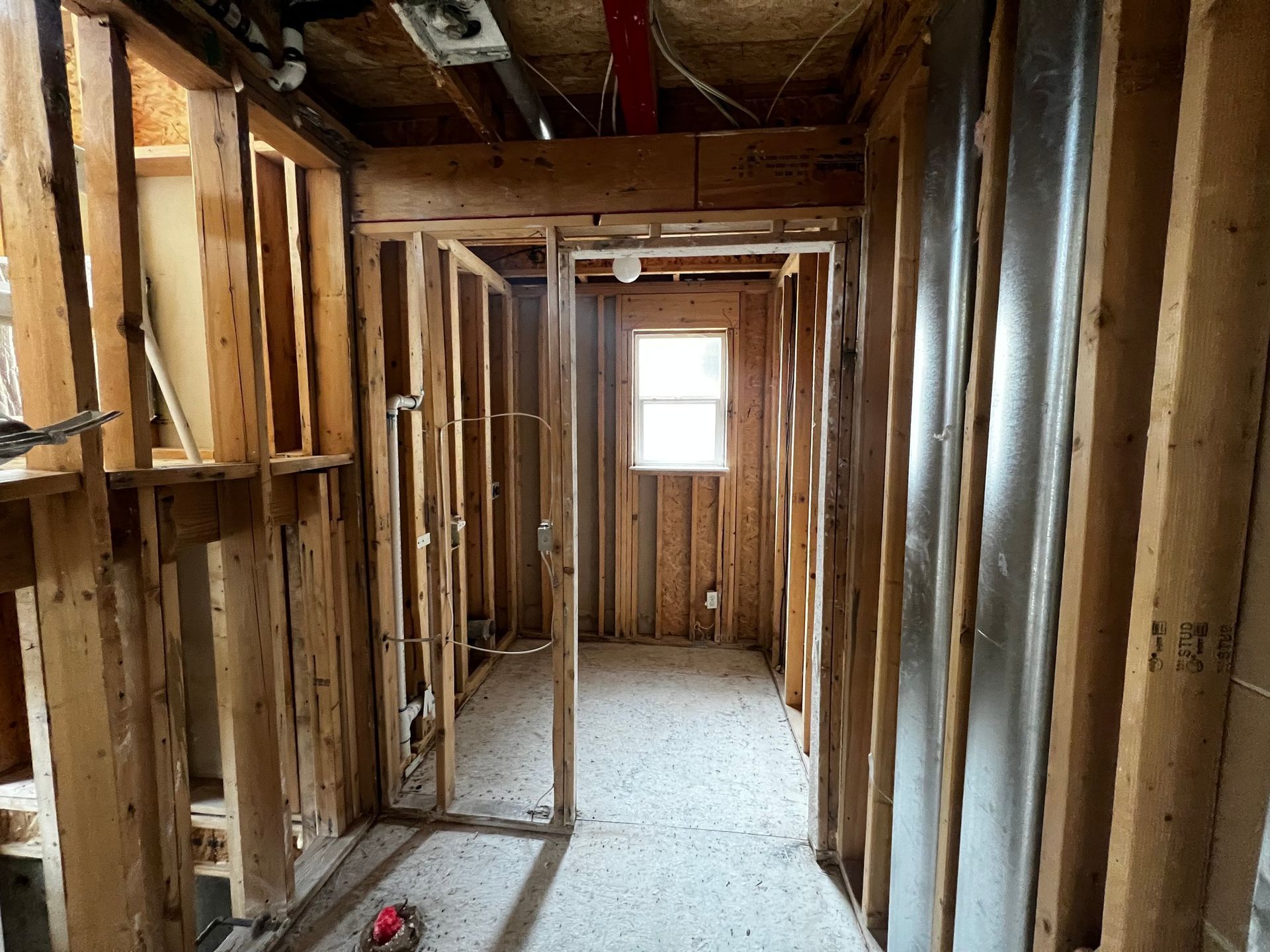 Construction site showing a narrow hallway with exposed wood framing, a subfloor, and a window at the end.