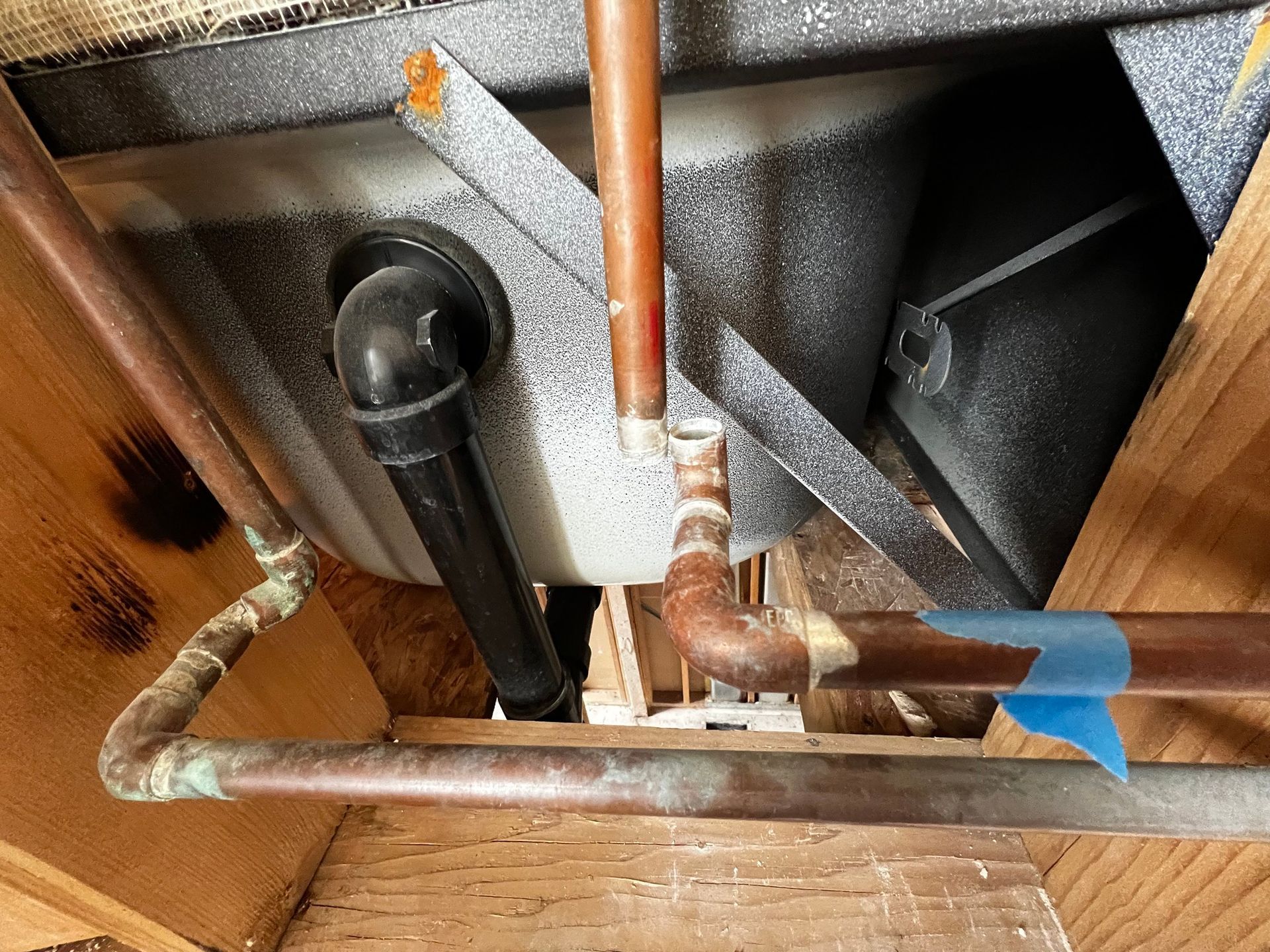 A high-angle view shows copper pipes and a black plumbing drain installed near a speckled gray tub in a wooden frame.