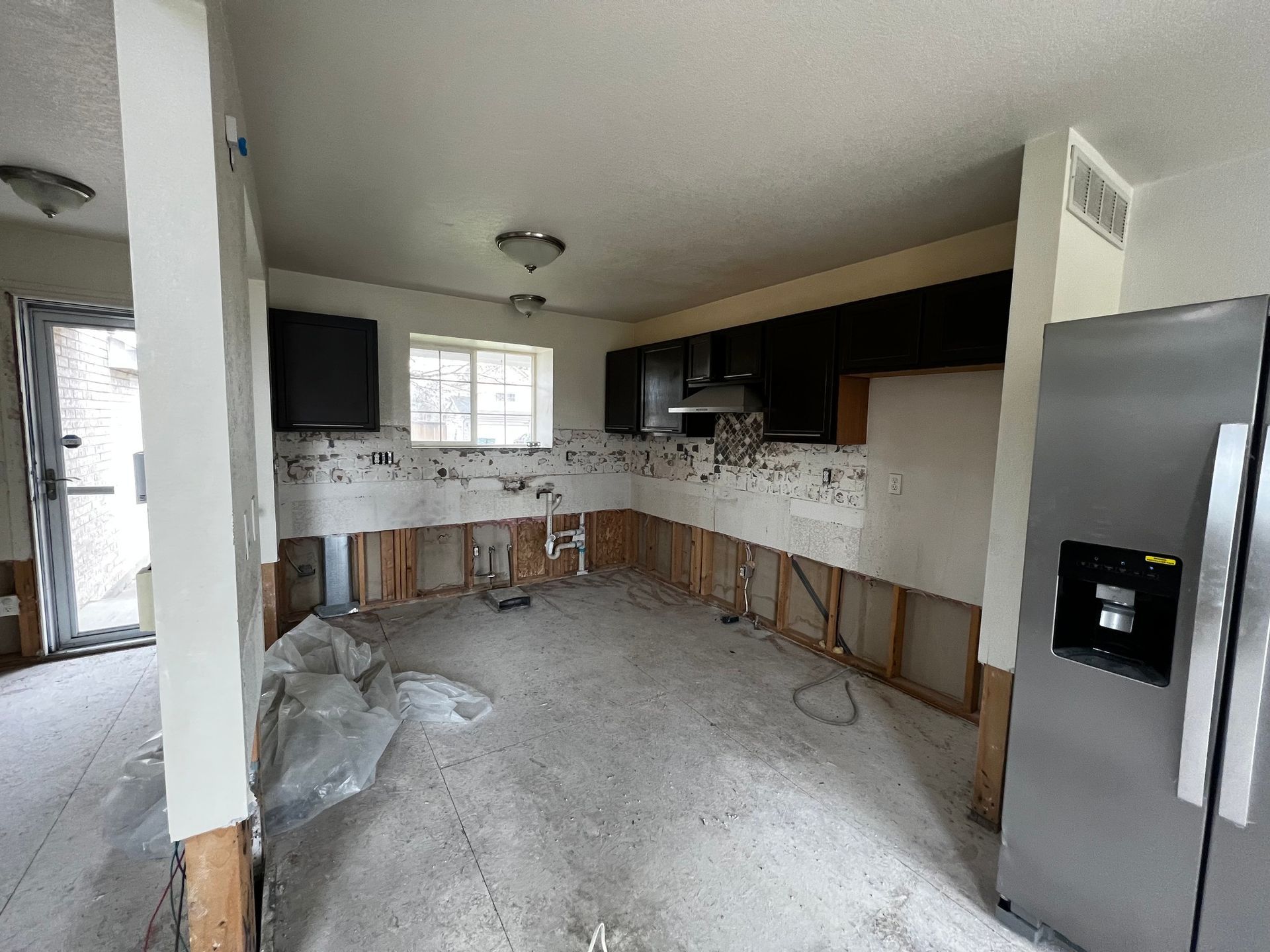 An unfinished kitchen renovation with exposed wall studs, dark upper cabinets, and a stainless steel refrigerator.