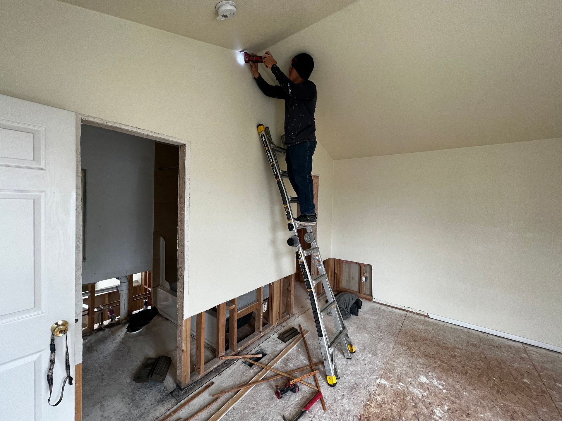A person stands on a tall, folded ladder, using a power drill on a wall corner in a room under construction.