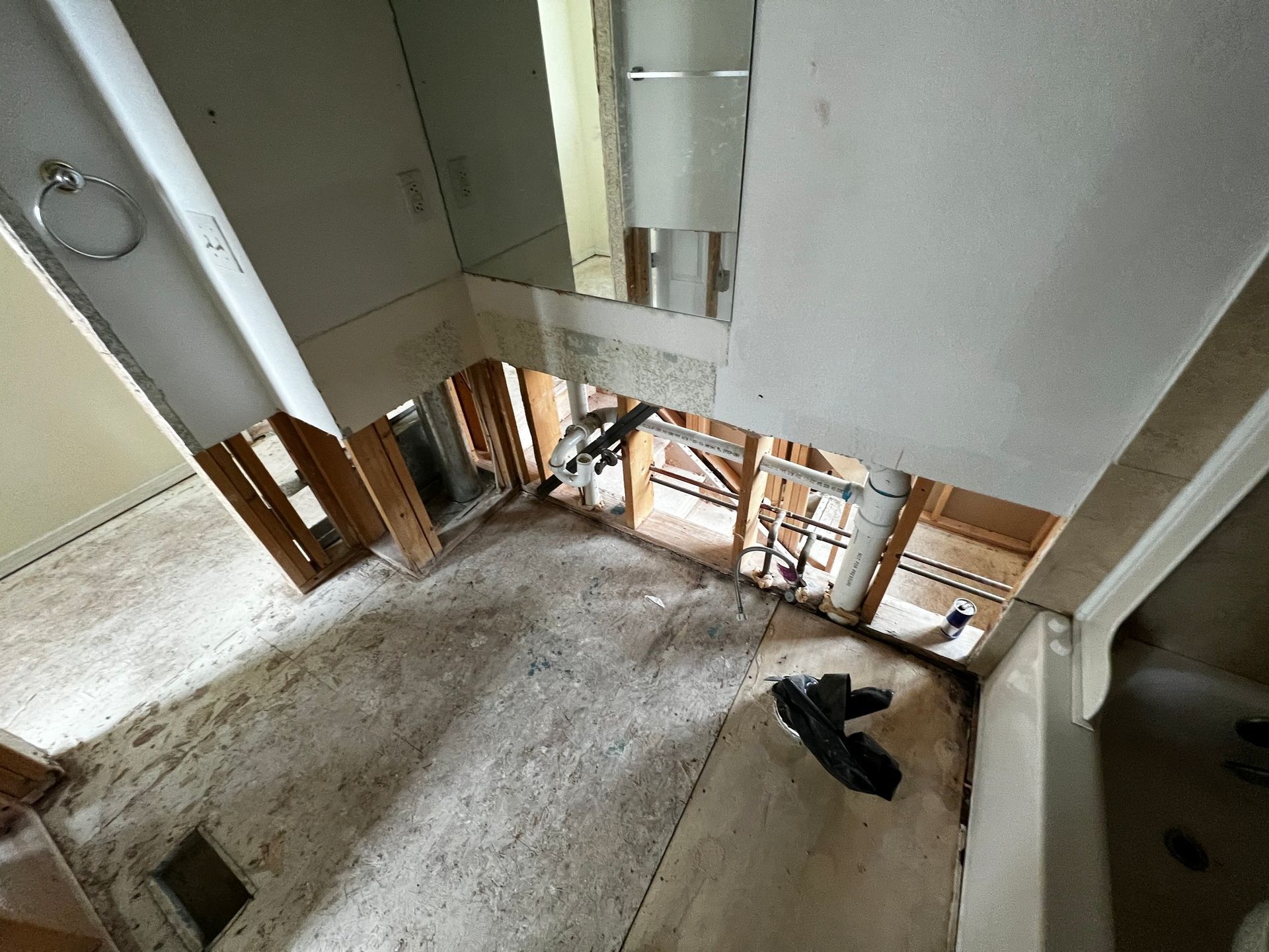 Bathroom undergoing renovation with drywall removed, exposing wall studs and plumbing pipes near a mirror and bathtub.