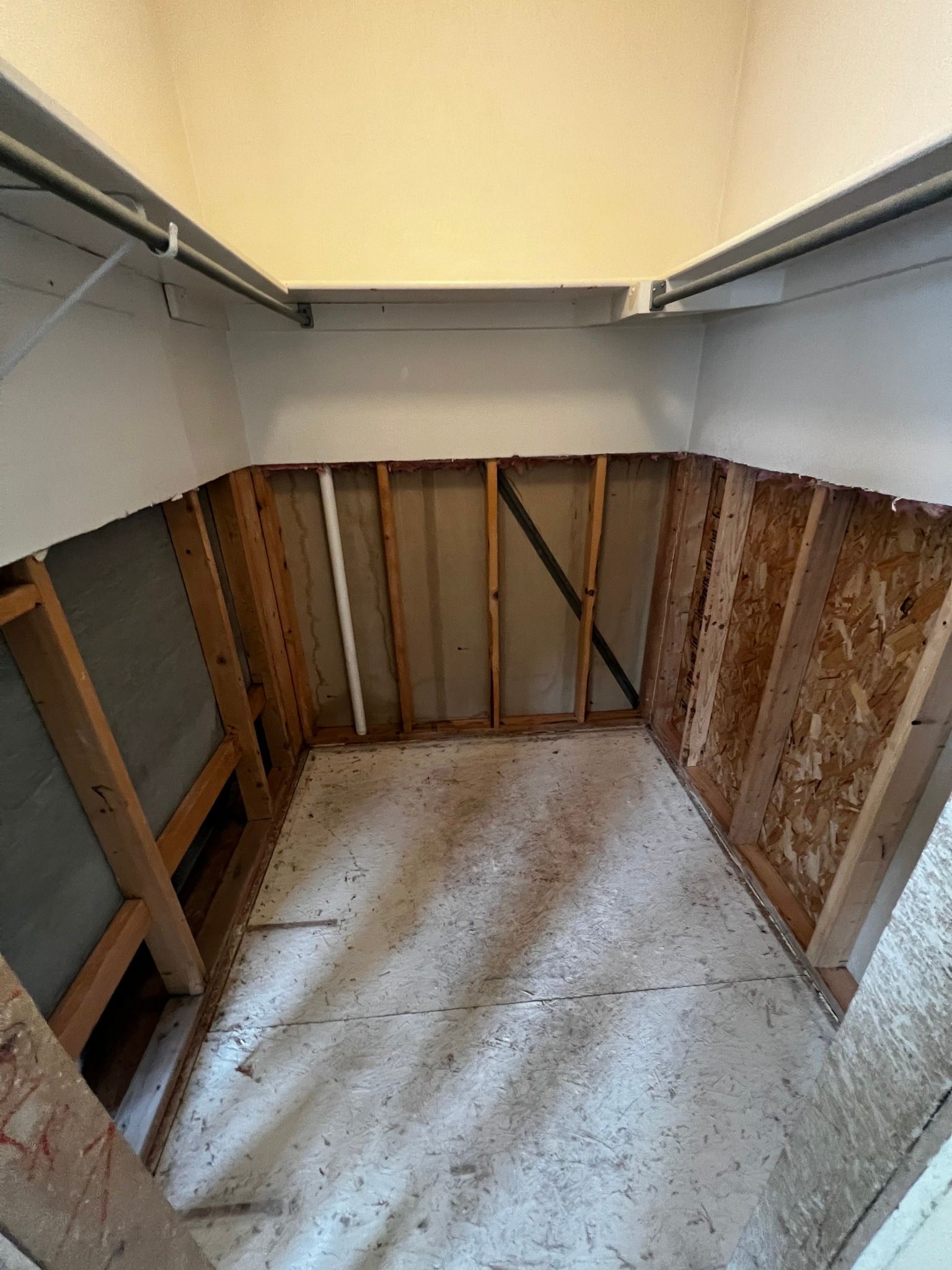 An empty closet with open wooden wall studs, exposed floor subflooring, and bare white upper walls.