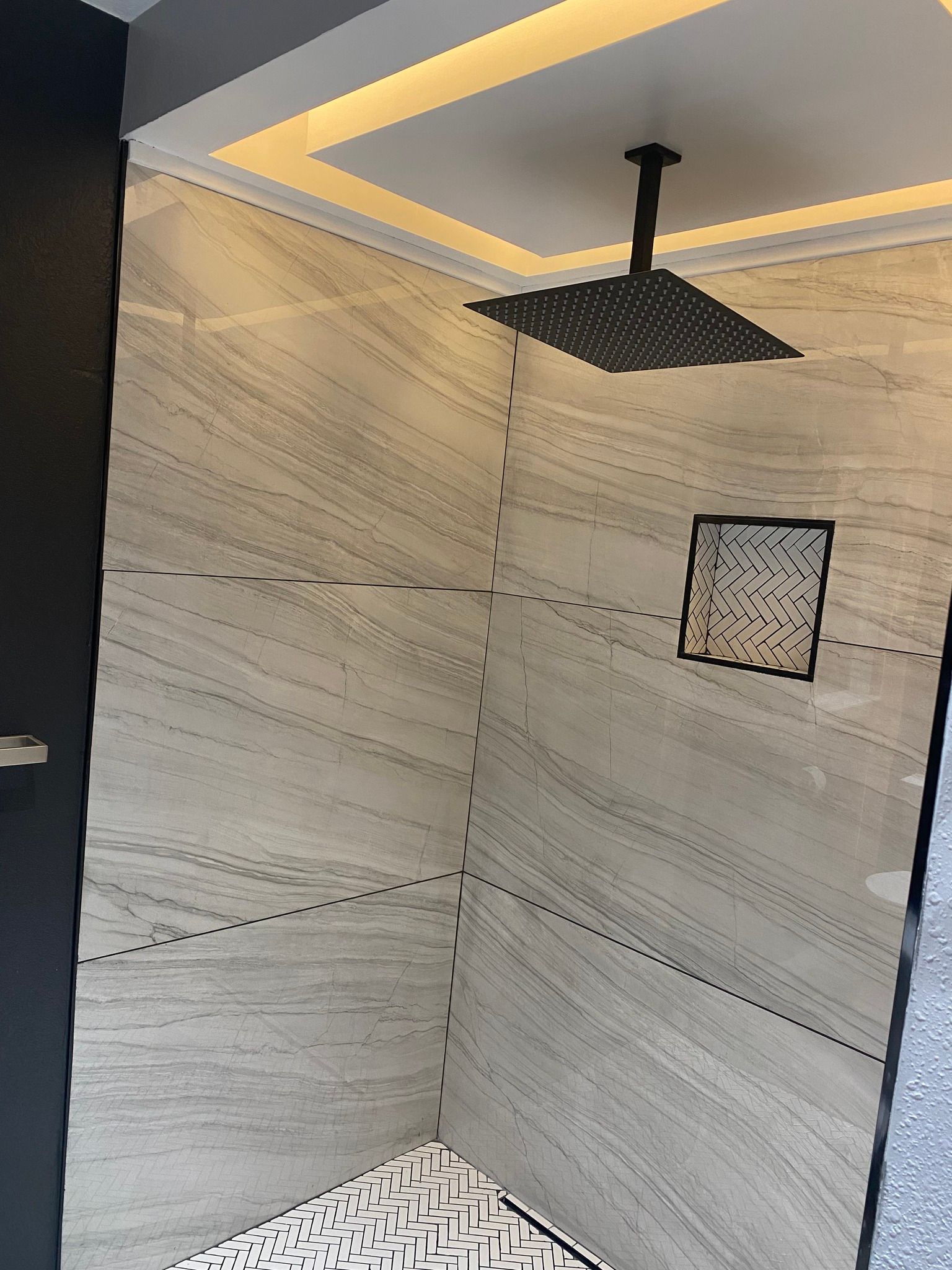 A modern walk-in shower with light beige stone-look tile walls, a matte black square rainfall head, and patterned floor.