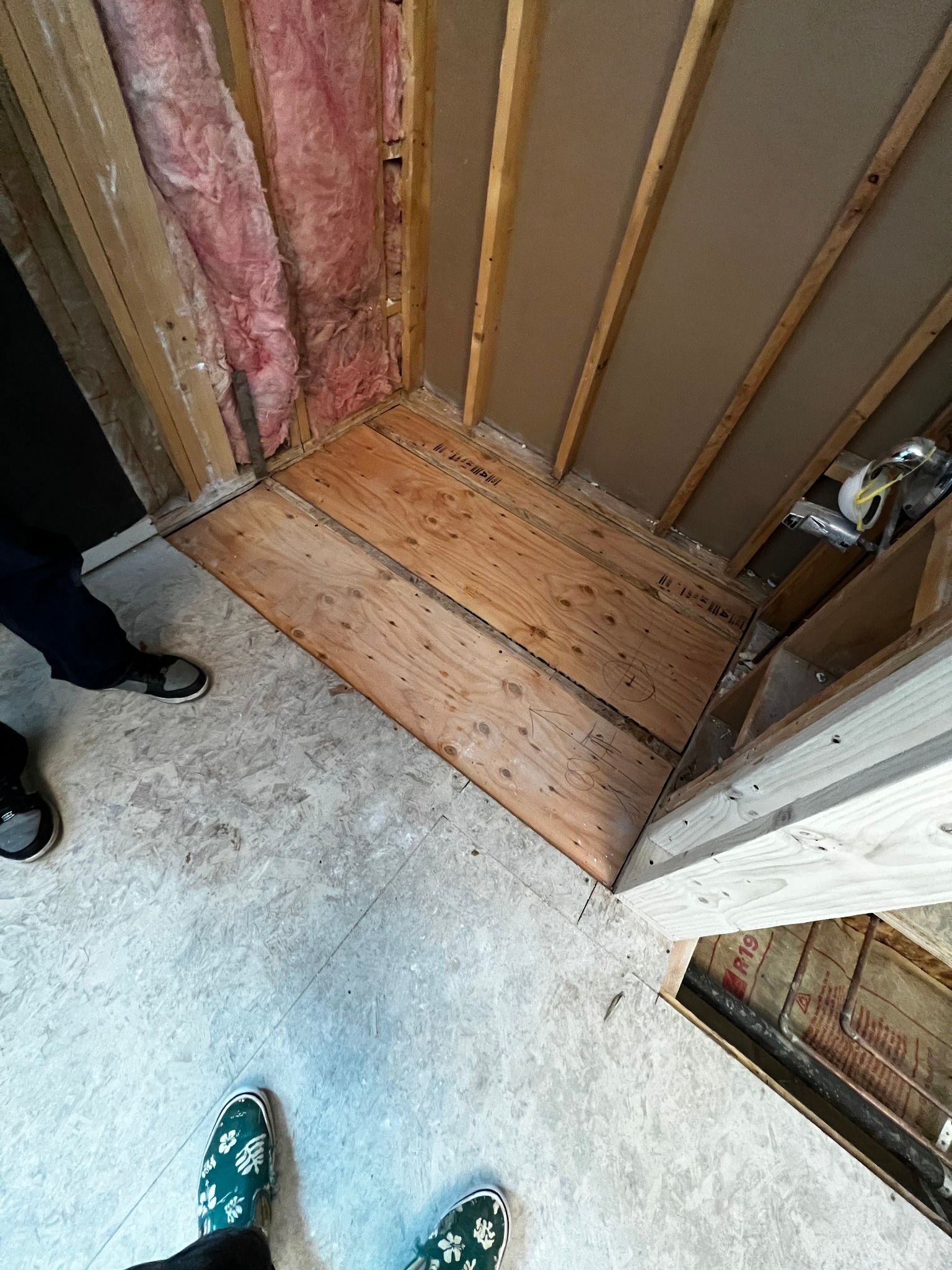 An unfinished shower stall showing exposed wooden studs, insulation, and a plywood base in a room under construction.