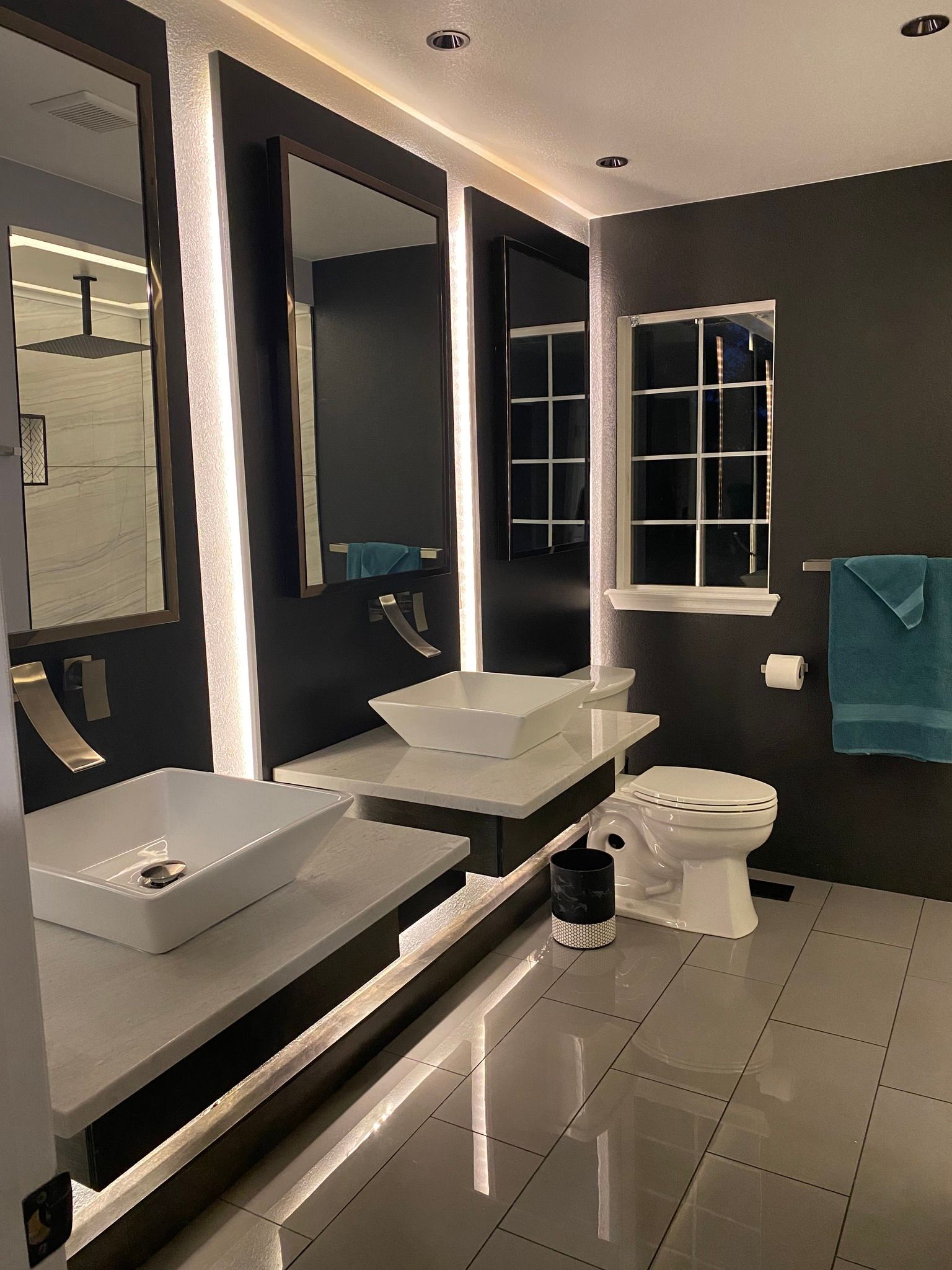 A modern bathroom with two white vessel sinks on a lit floating vanity, dark walls, a window, and a toilet.