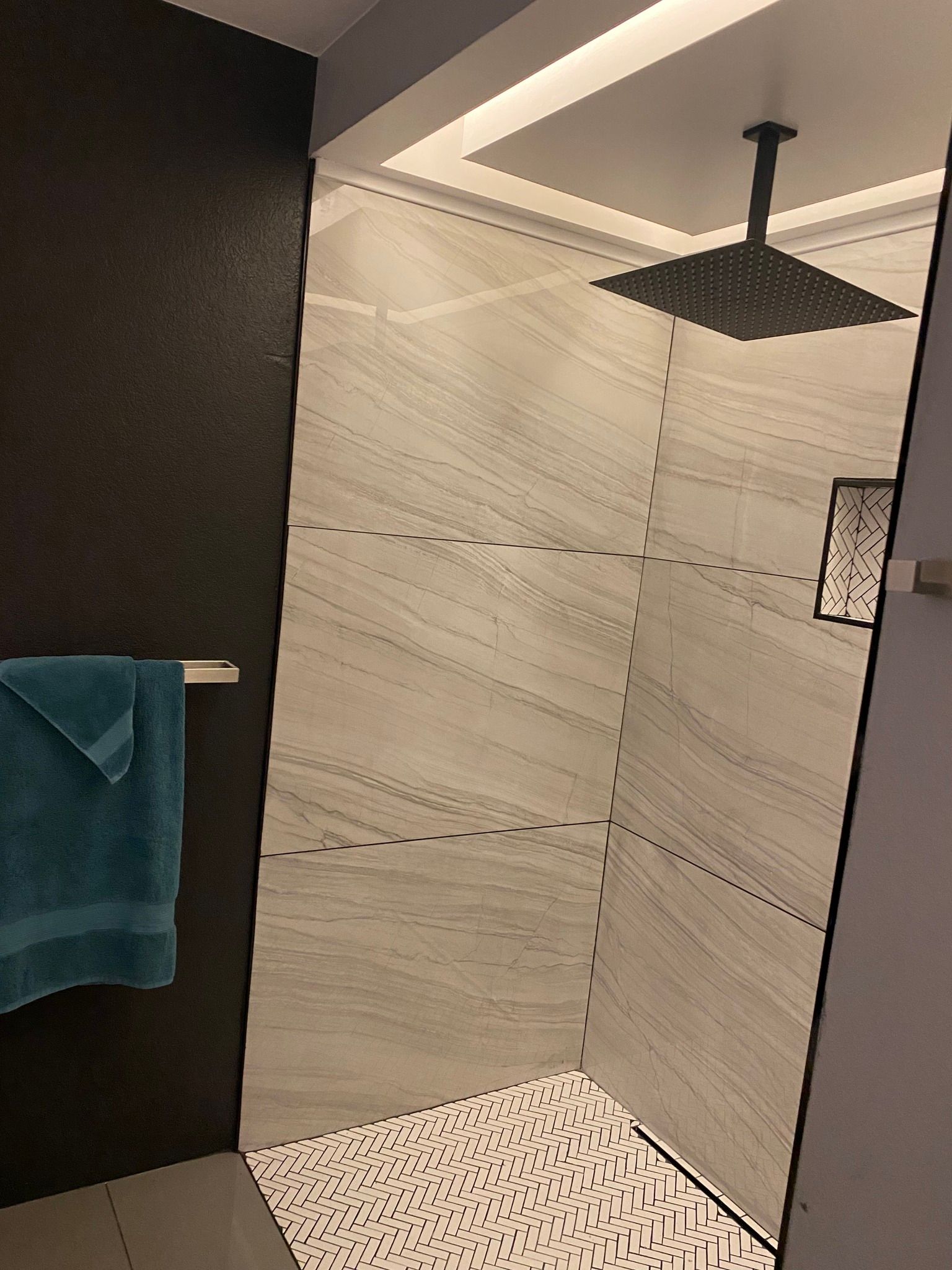 A modern shower with light-veined tile walls, a patterned tile floor, a square overhead rainfall head, and a blue towel.