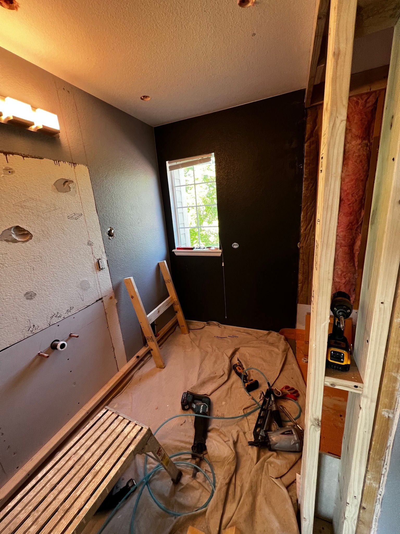 Interior of a bathroom under renovation with exposed wood framing, drywall patches, tools, and a window on a dark wall.