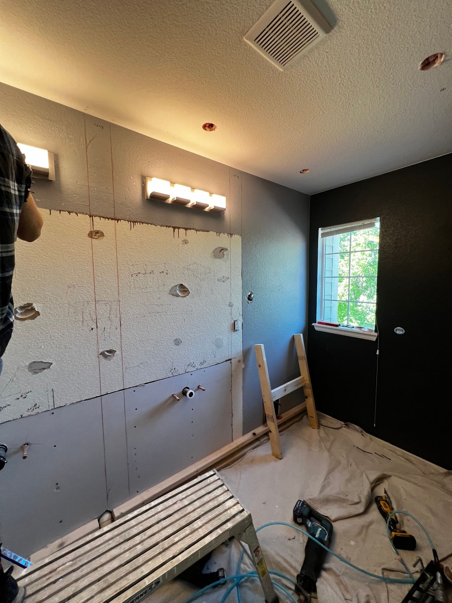 A person works on a bathroom renovation, installing tile backer board on a grey wall beneath a bright vanity light.