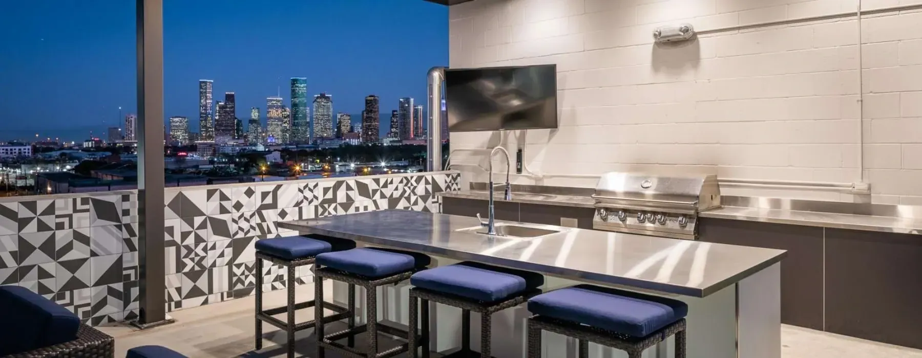Rooftop outdoor kitchen with bar seating and a grill, overlooking a city skyline at dusk.