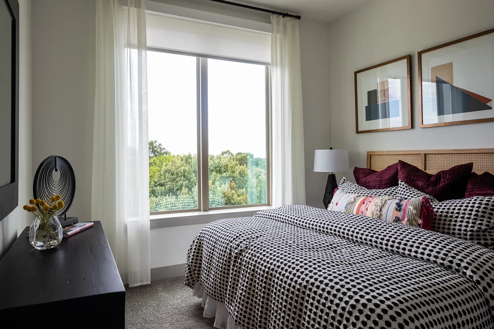 Bedroom with a large window overlooking trees, a black dresser, and a bed with patterned bedding.