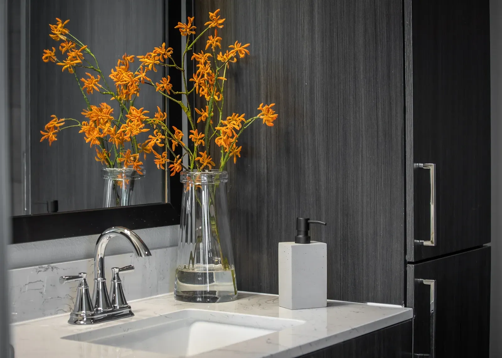 Modern bathroom vanity with chrome faucet, white countertop, and a vase of orange flowers.