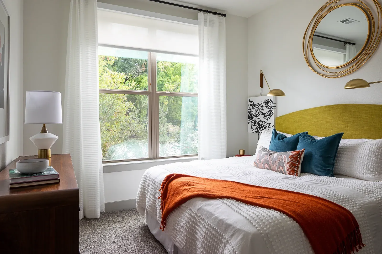 Bedroom with large window overlooking trees and water, featuring a king-size bed with orange and blue accents, and a gold circular mirror above the headboard.