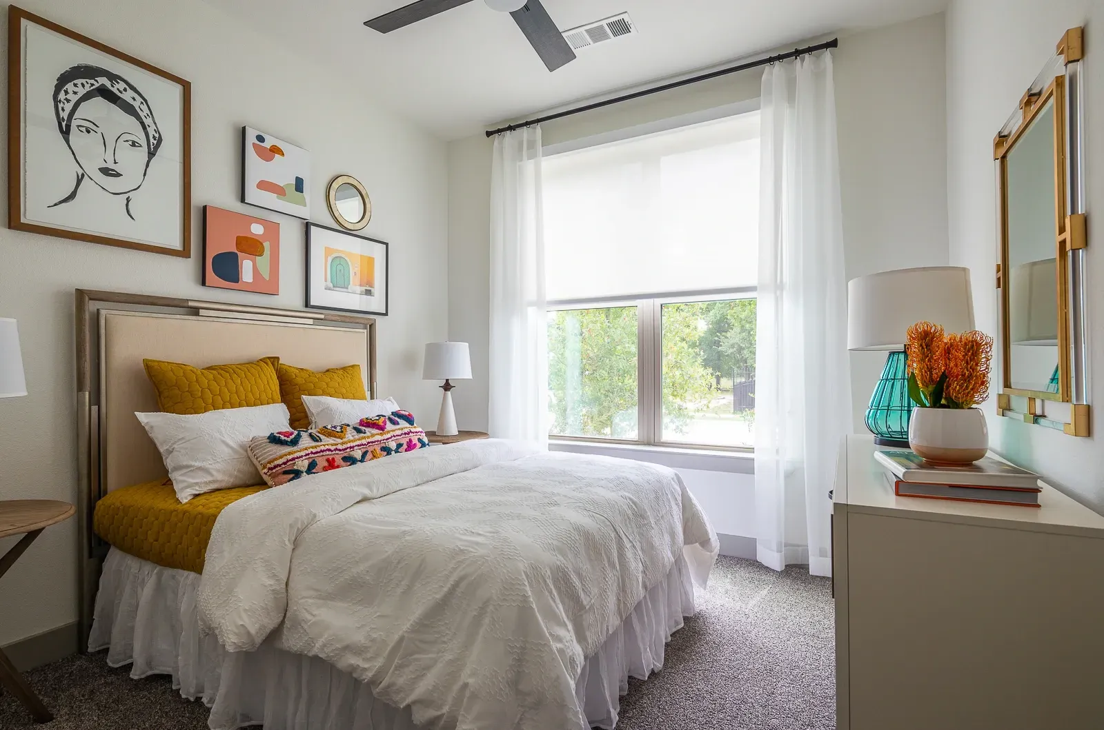 Cozy bedroom with a queen-size bed, plush bedding, and decorative pillows. Features artwork on the wall and a large window with white curtains.